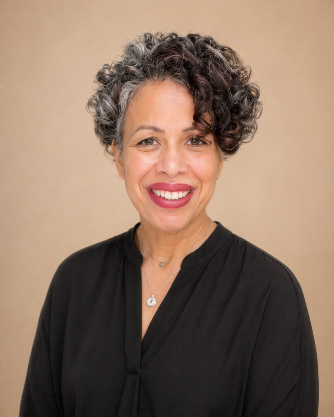 A woman with short curly gray hair, wearing a black sleeveless top, sitting on the floor in a living room, smiling at the camera, with her hands clasped in front of her.
