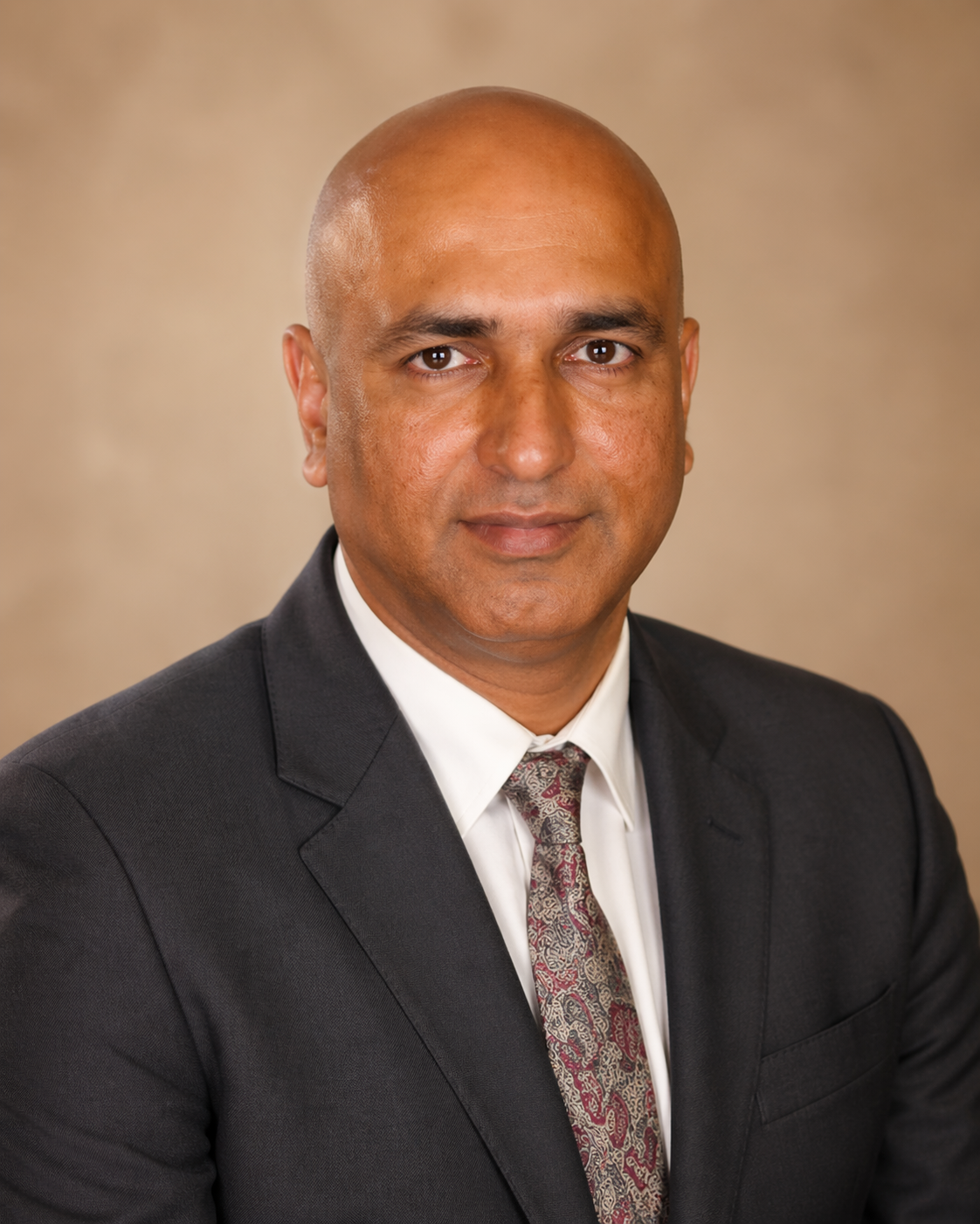 A bald man wearing a dark suit, white shirt, and patterned tie, sitting in a chair with a decorative gold background.