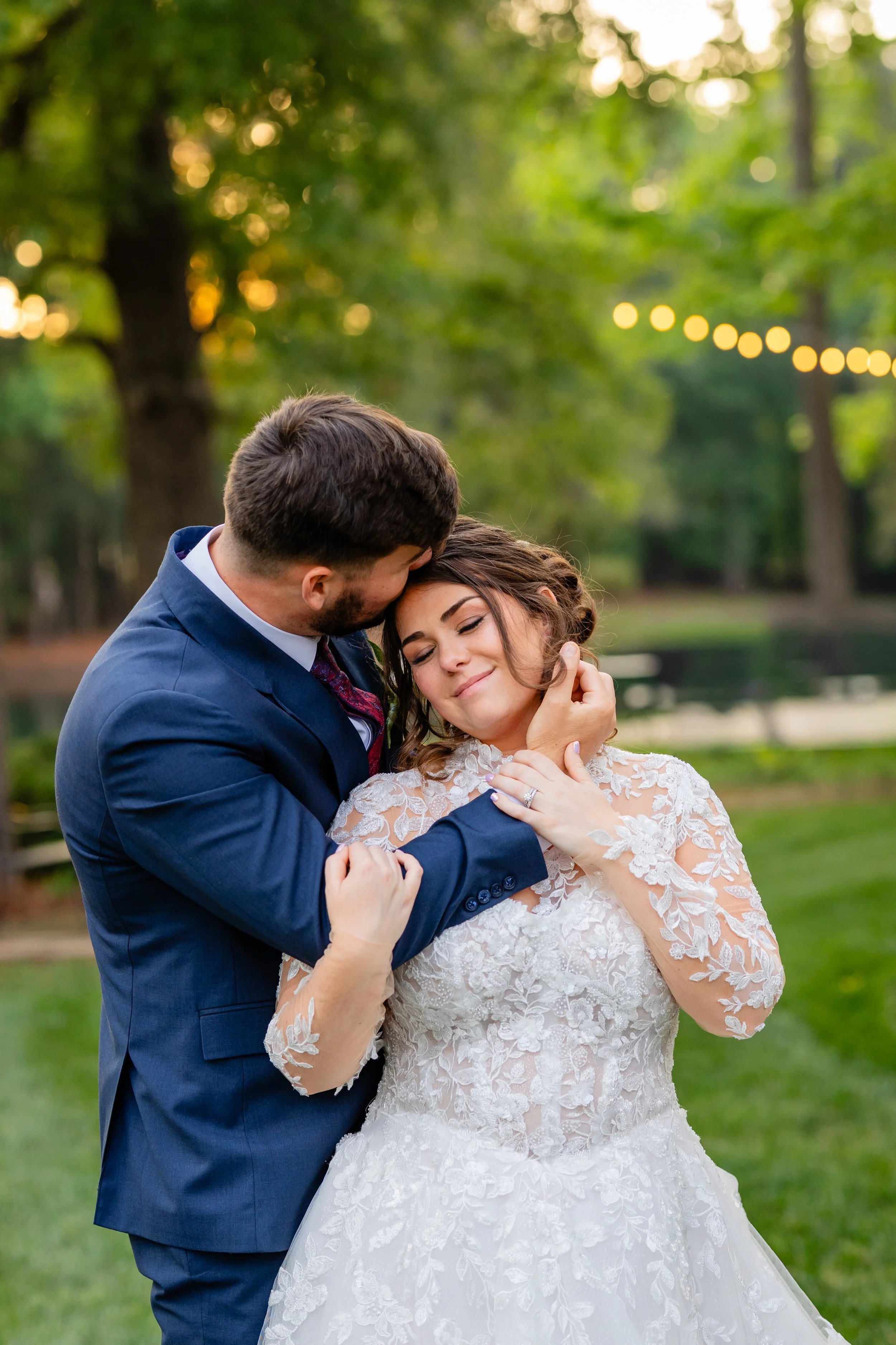 A bride and groom embrace outdoors during sunset. The groom wears a blue suit, and the bride wears a lace wedding dress. They hold each other tenderly with a blurred background of green trees and string lights.