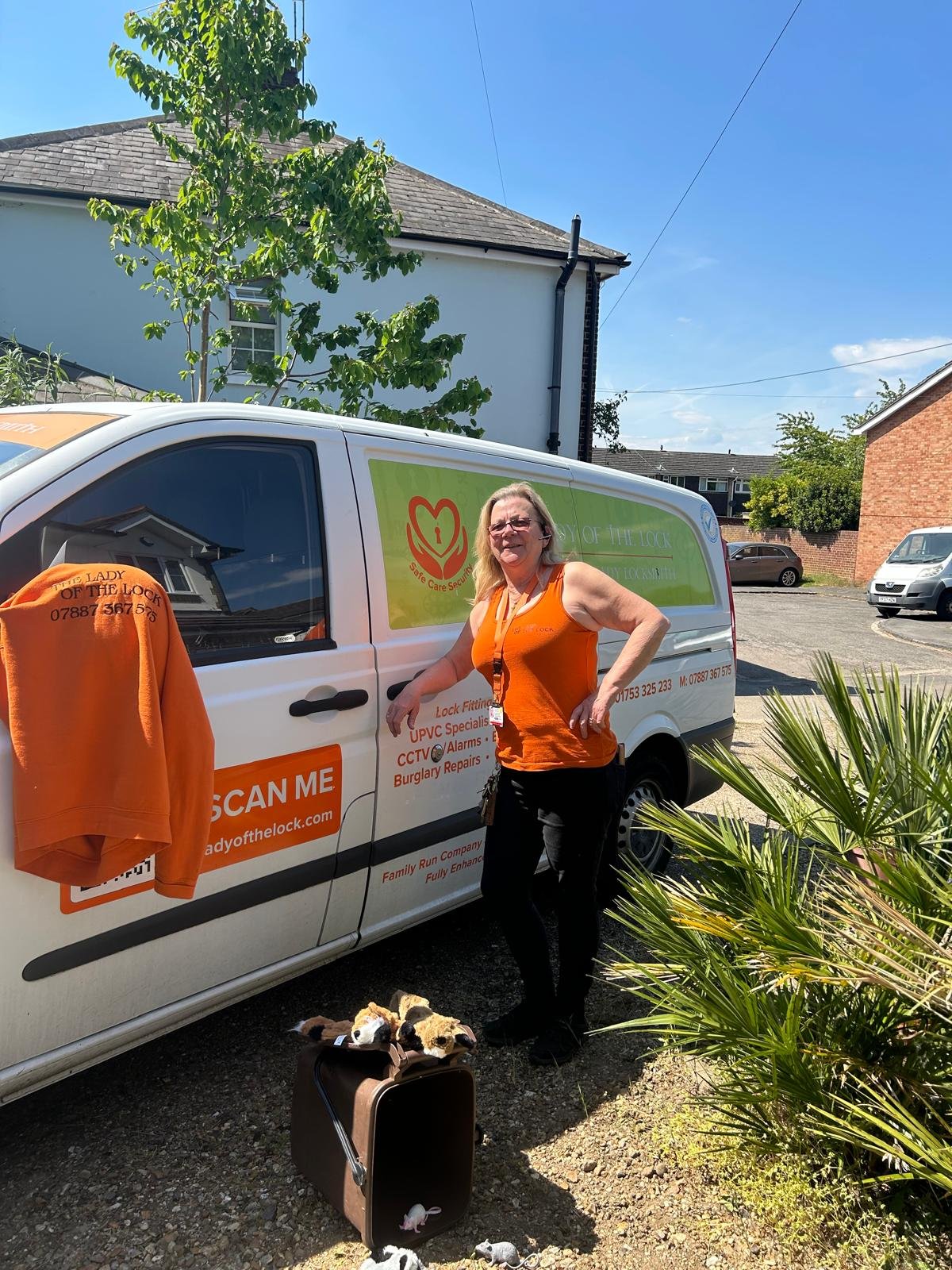 A woman standing next to a branded service van labeled 'Lady of the Lock' with a phone number and website. She is wearing an orange tank top and has a set of keys around her neck. On the ground are a brown pet carrier with plush animal toys and some