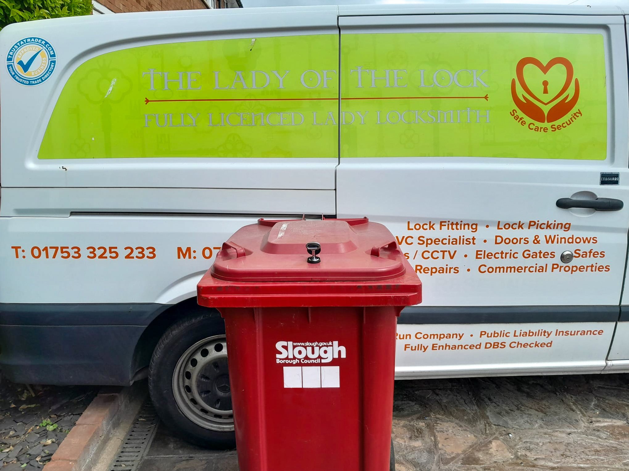 A white van with green and orange signage advertising locksmith services, parked next to a red waste bin marked 'Slough Borough Council' on a stone pavement.