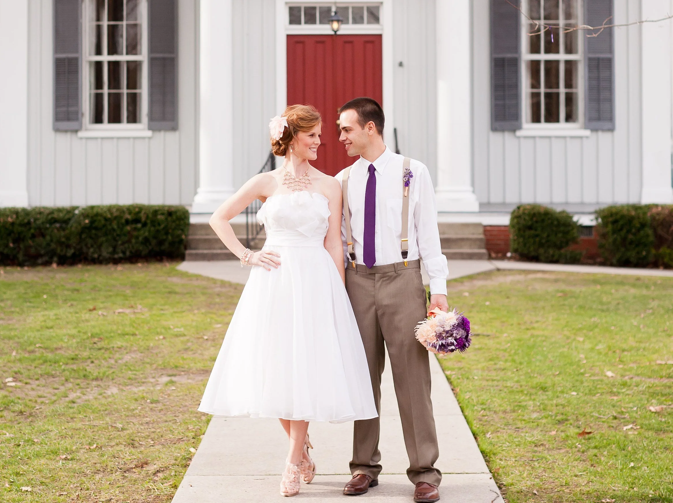 A bride and groom walking on a sidewalk in front of a white building, smiling and looking at each other. The bride is wearing a white dress with a flower in her hair, and the groom is in a white shirt with suspenders and holding a flower bouquet.