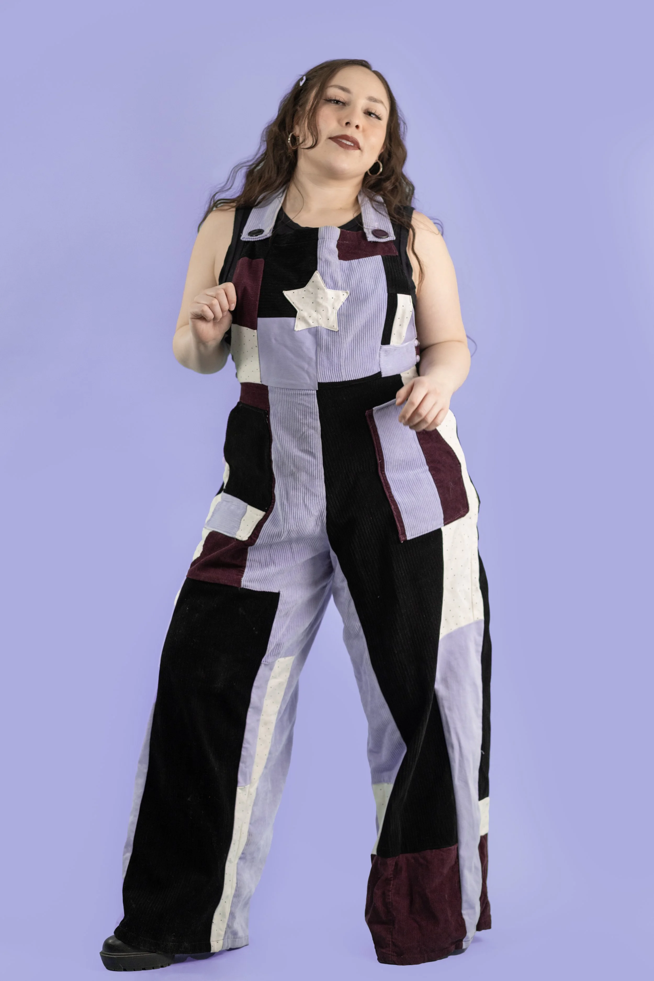 A woman with long, wavy brown hair and light skin, wearing dark lipstick, posing against a purple background. She is dressed in a patchwork jumpsuit with various colors including black, purple, white, and maroon, and accessories with hoop earrings.