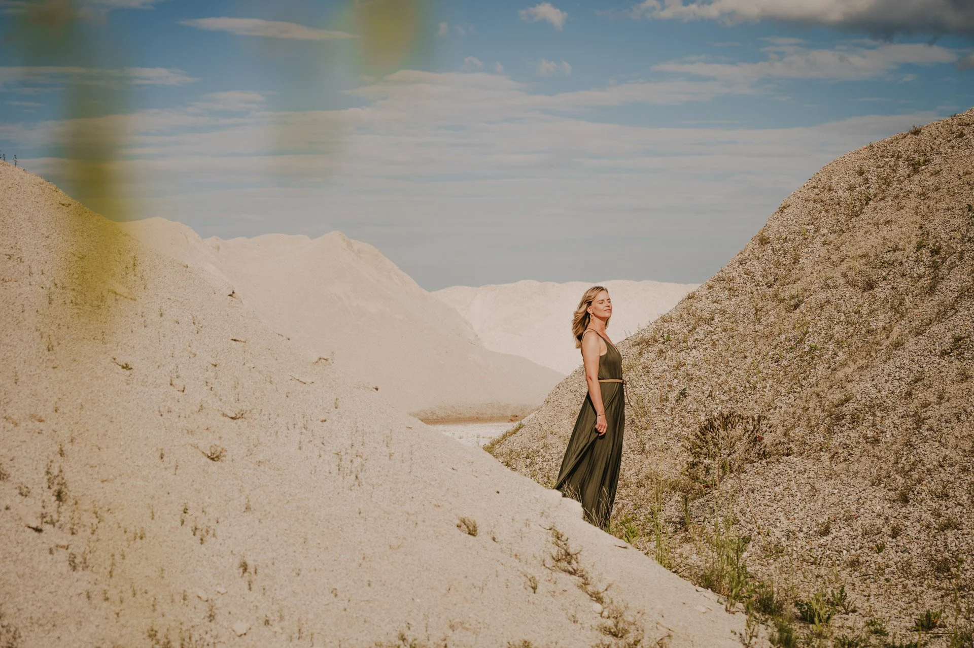 Frau in einem langen, grünen Kleid steht zwischen sandigen Hügeln in einer wüstenähnlichen Landschaft mit blauem Himmel.