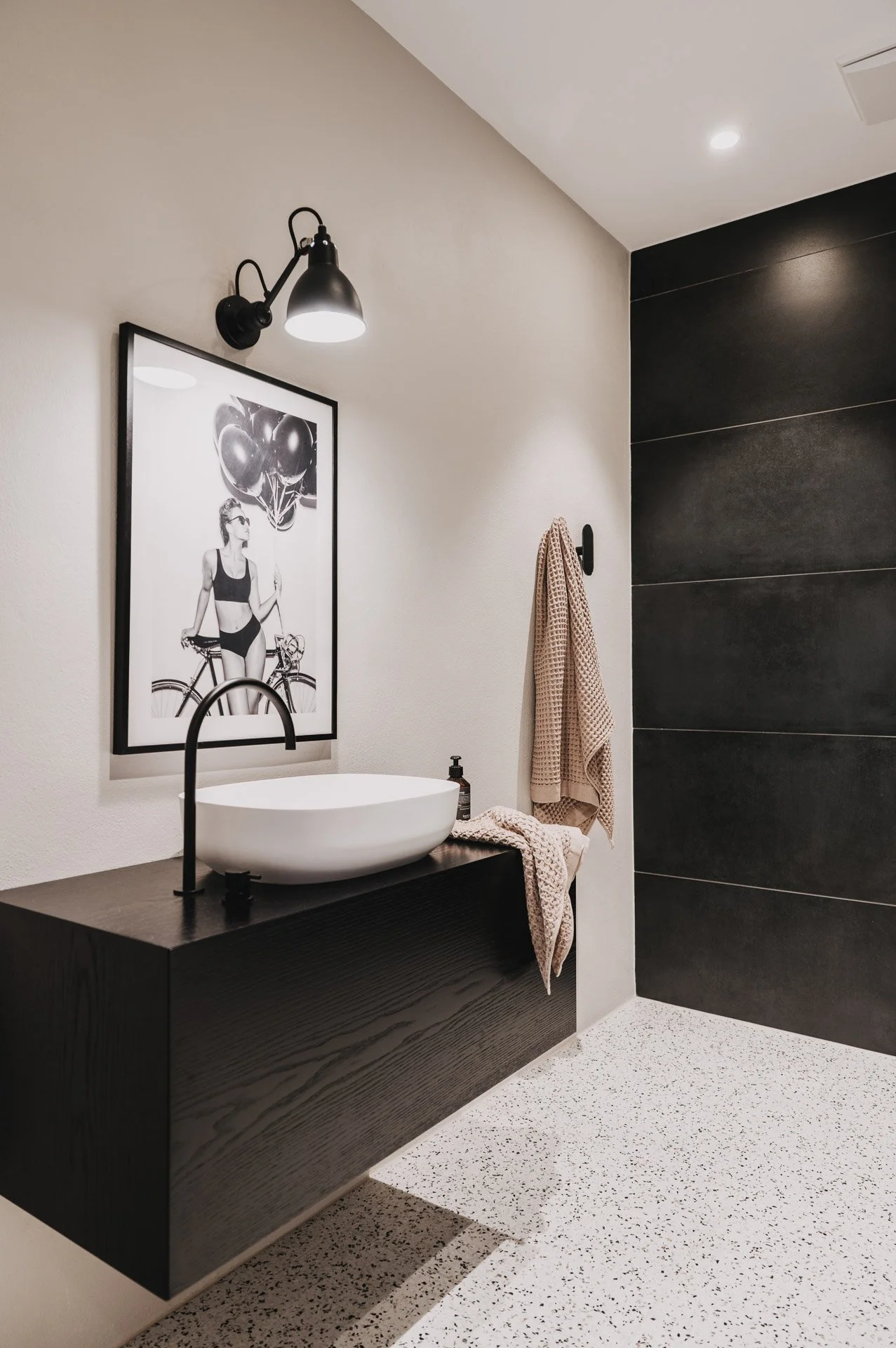 Badezimmer mit modernem Design, schwarzen Fliesen, einem Waschbecken auf einem schwarzen Unterschrank, einem Handtuch, einem Seifenspender und einem gerahmten Bild an der Wand.