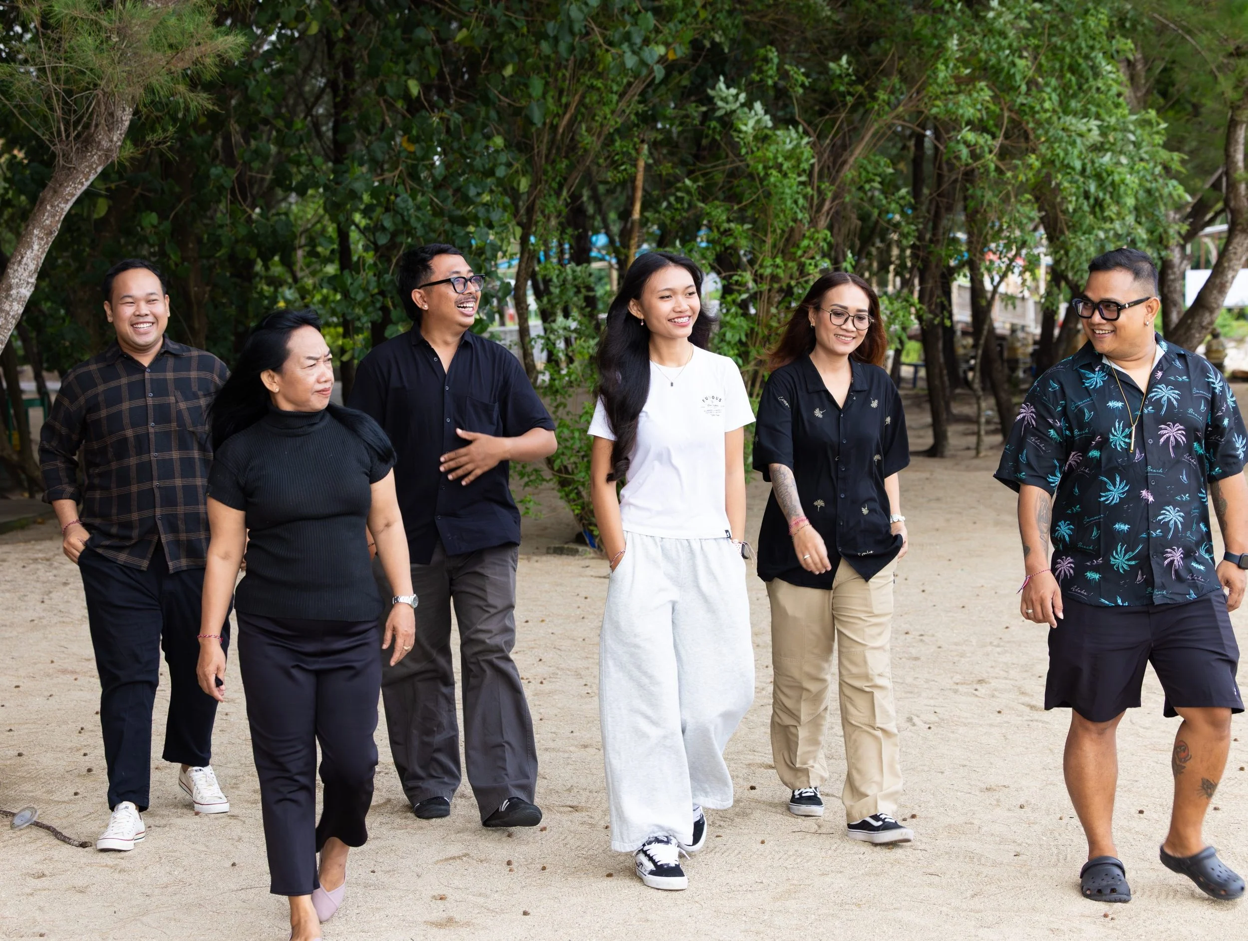 Group of seven diverse people walking and smiling outdoors on a dirt path surrounded by green trees.