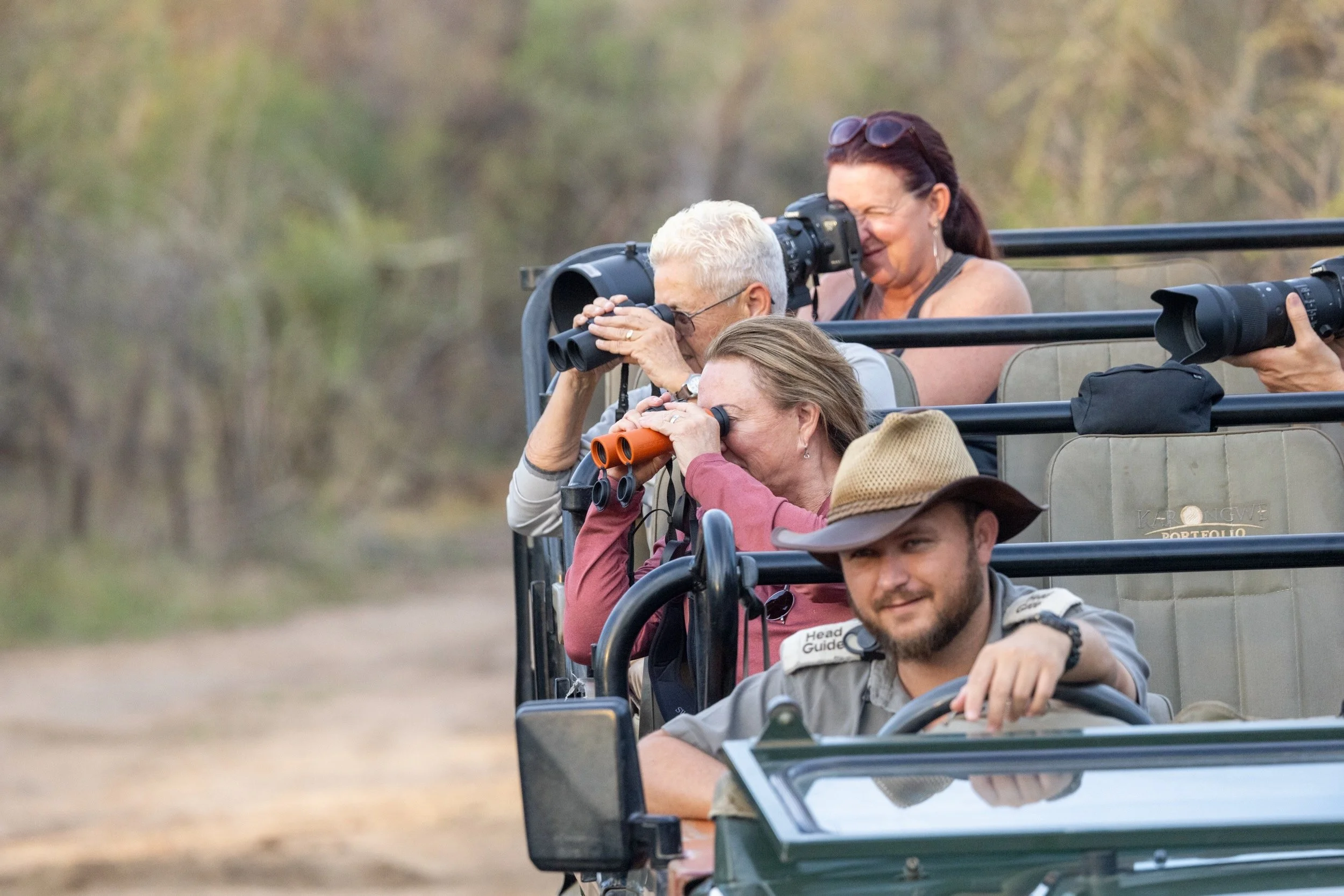 Four people on an open-air safari vehicle, observing wildlife through binoculars and cameras, in a natural outdoor setting.