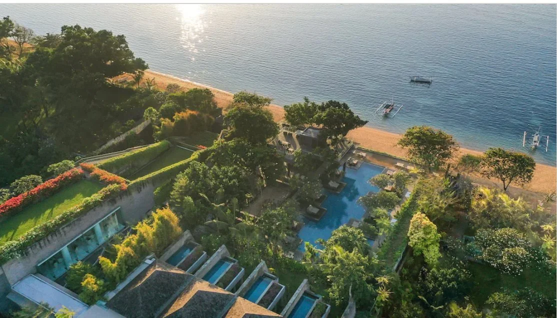 Aerial view of a beachfront resort with a large swimming pool surrounded by lush green trees and gardens. The resort is near a sandy beach with calm water and small boats anchored offshore.