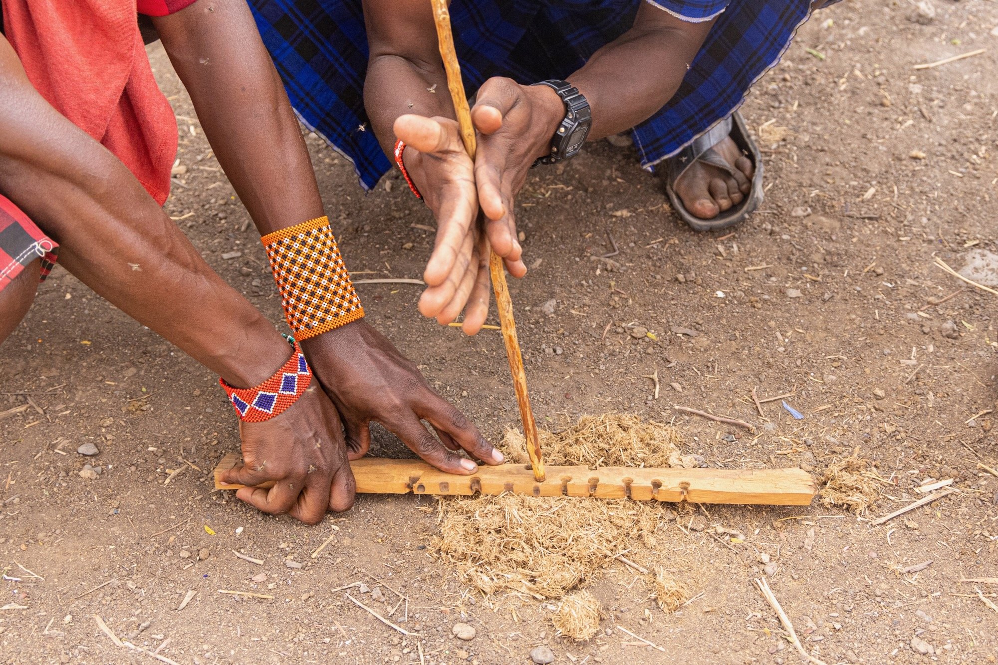 Two people working together to cut dried grass with a wooden tool on the ground, wearing traditional beaded jewelry and casual clothing.