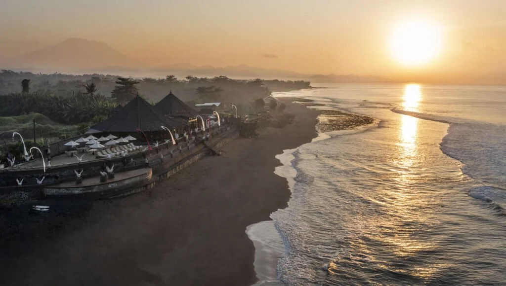 Sunset over a beach with waves lapping the shore, and a row of black buildings and umbrellas along the sand.