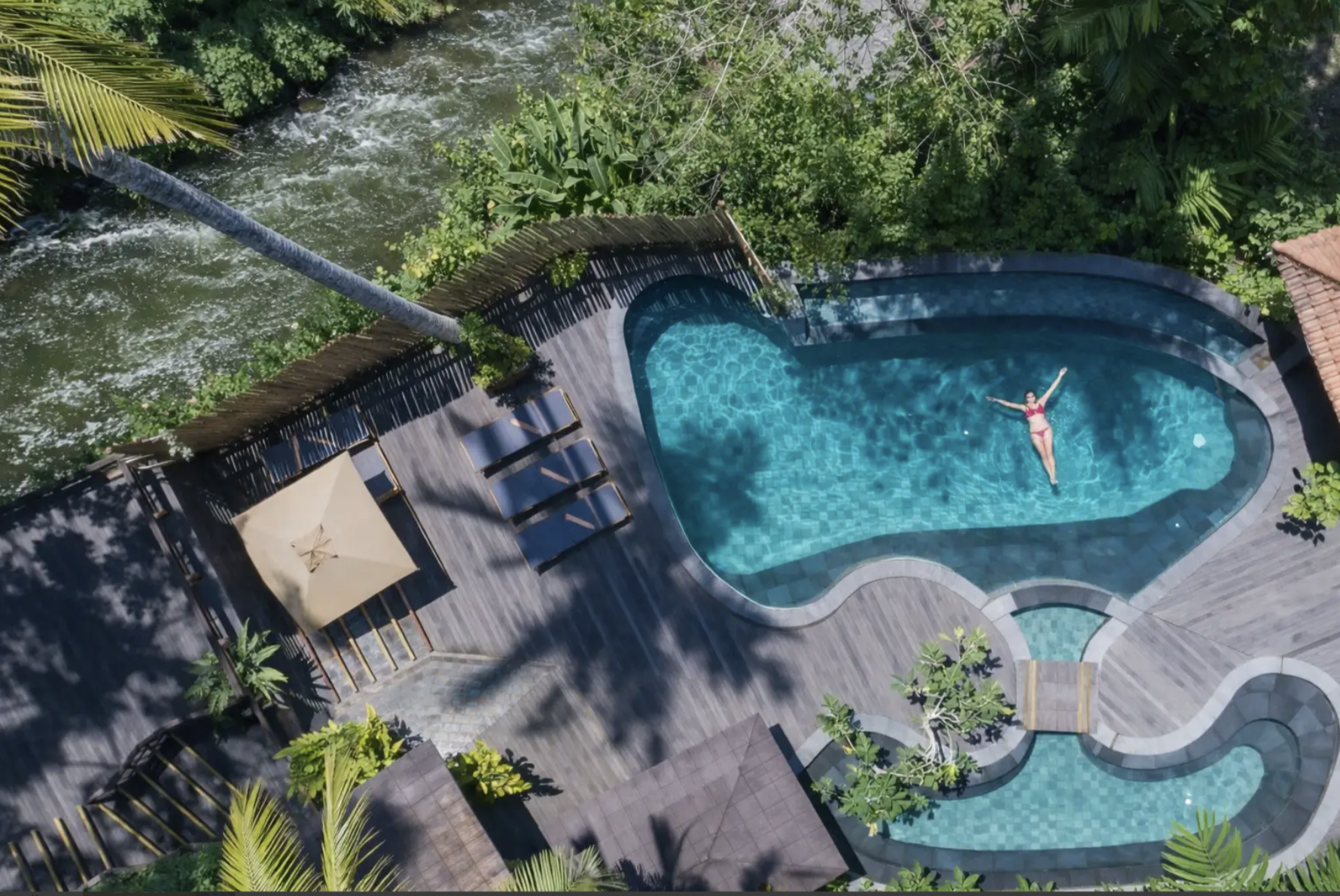 Aerial view of a swimming pool area with a woman in a red swimsuit floating on her back, surrounded by lush green trees and wooden deck chairs.