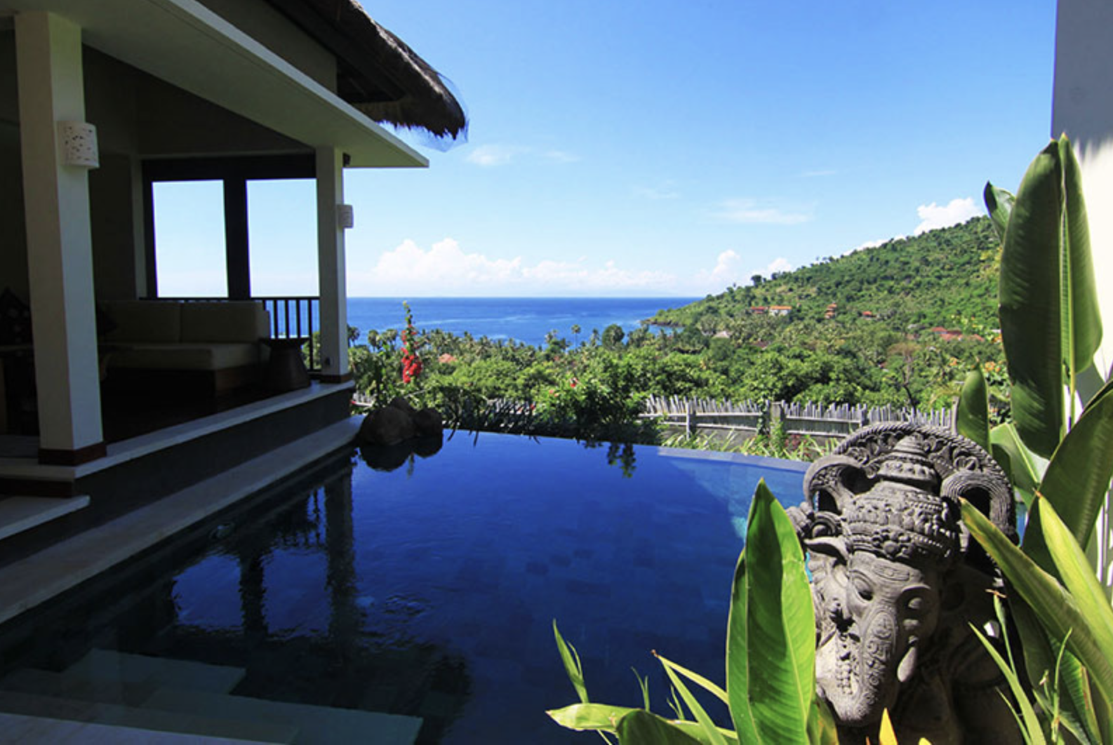 A tropical resort view featuring a private pool overlooking lush green hills and the ocean, with a decorative stone sculpture and tropical plants in the foreground.