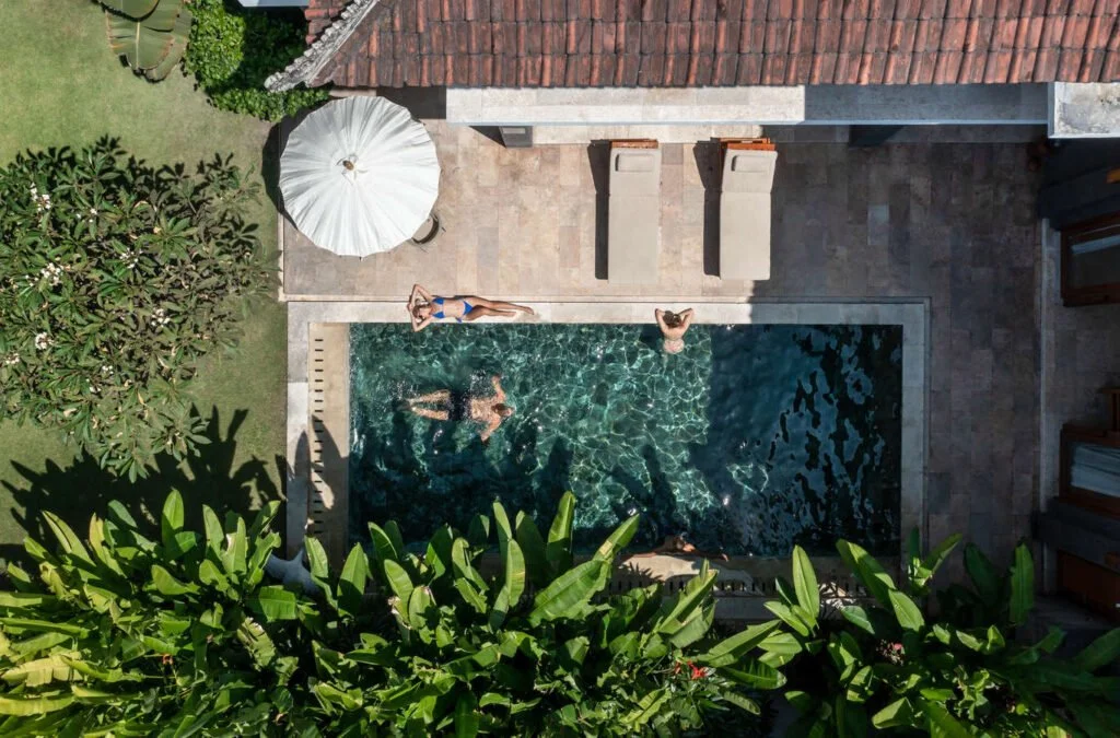 Top-down view of a backyard with a swimming pool, two people swimming, and one person lounging on the pool's edge. There is a large white patio umbrella and some lounge chairs near the pool. Lush green foliage surrounds the area, with parts of a building with a textured roof visible.