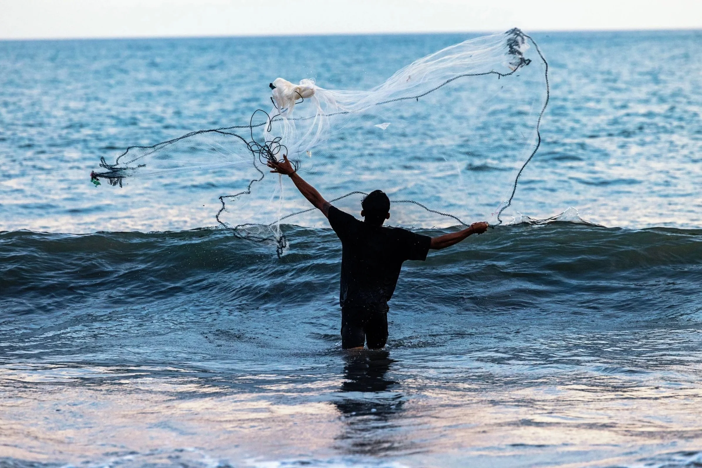 A person standing in the ocean water, throwing a fishing net with the ocean and blue sky in the background.
