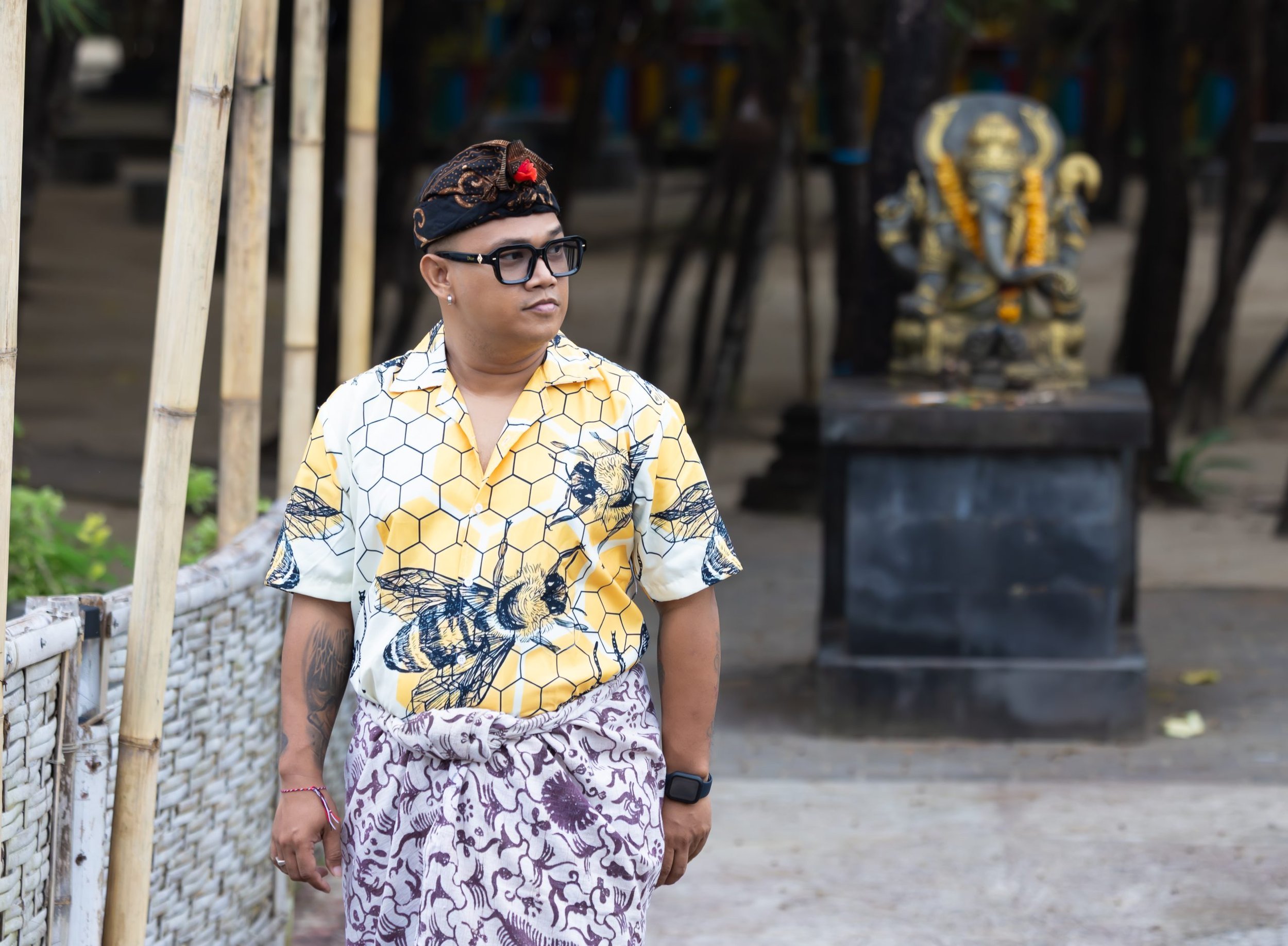 Man wearing glasses, a traditional Balinese headpiece, a yellow shirt with a honeycomb pattern, and patterned purple and white sarong standing outdoors near bamboo and a statue.
