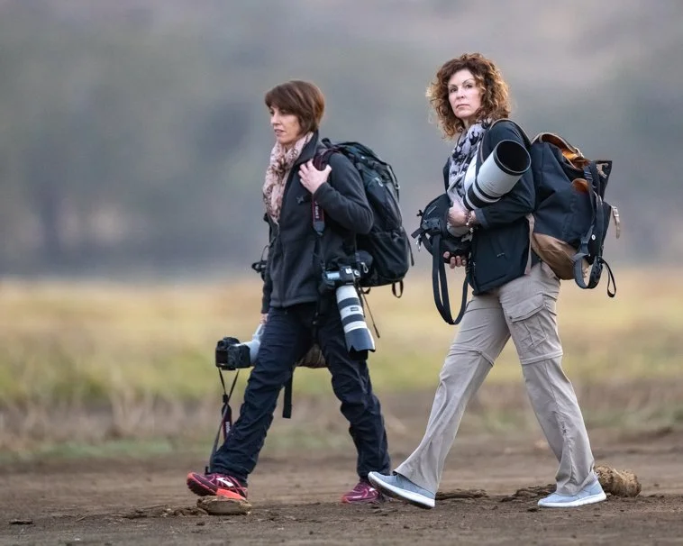 Two women carrying backpacks and camera gear walking outdoors on a trail.