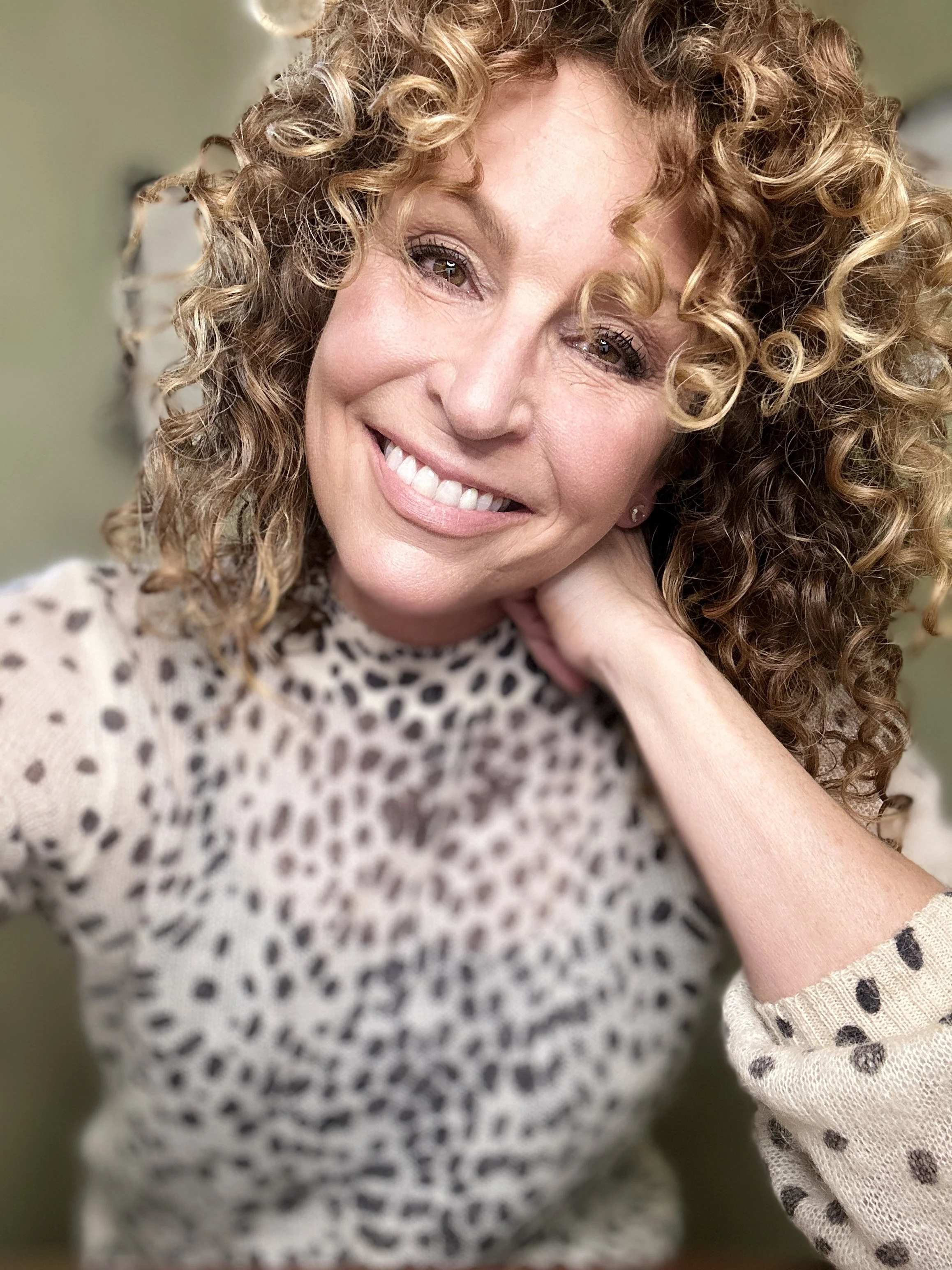 A woman with curly blonde hair wearing a leopard print top, smiling and resting her head on her hand.