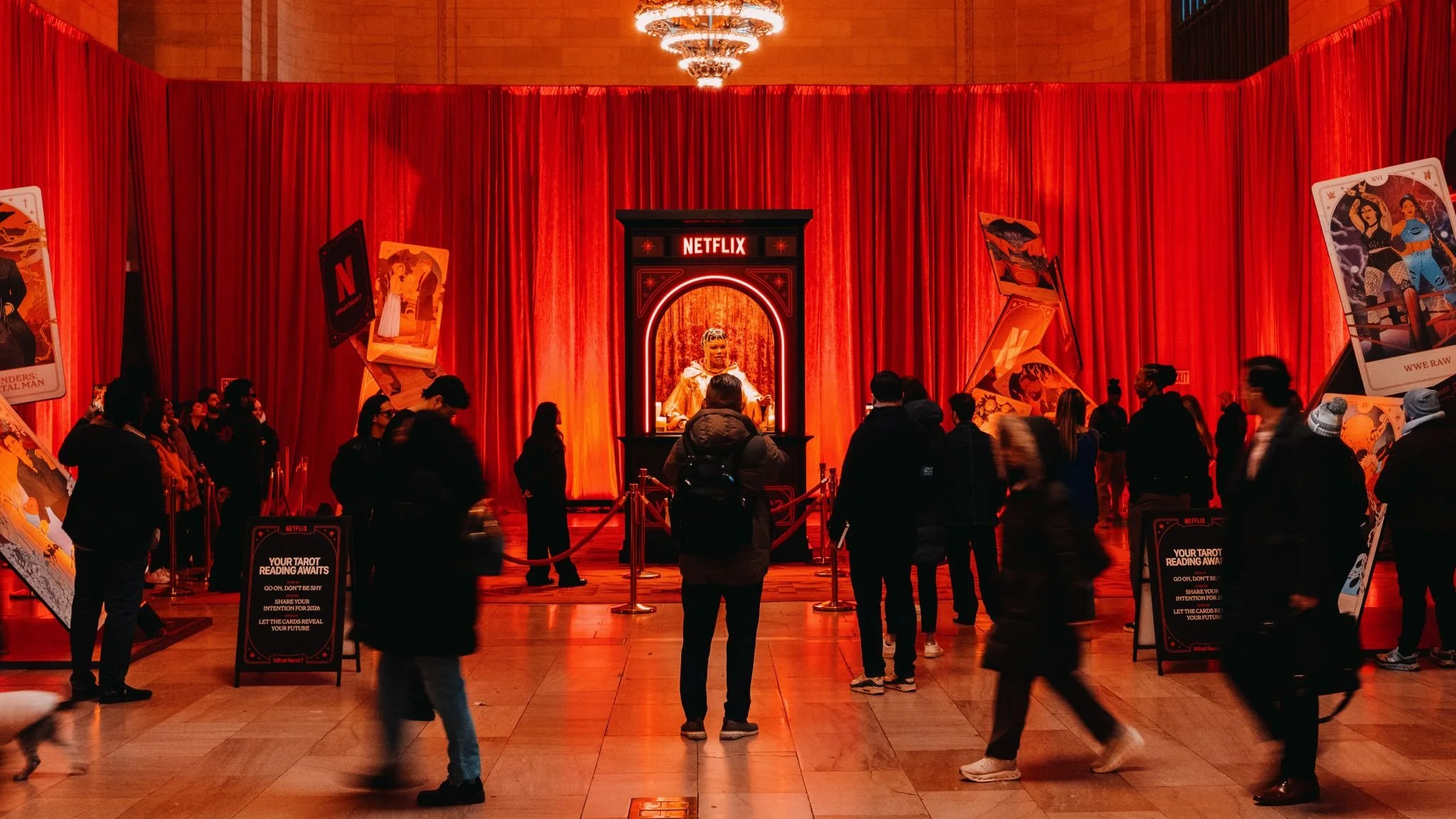 People gathered in front of a large red curtain with a central display featuring a figure and a Netflix sign. There are oversized playing cards and promotional signs for tarot reading and other entertainment on either side of the display.