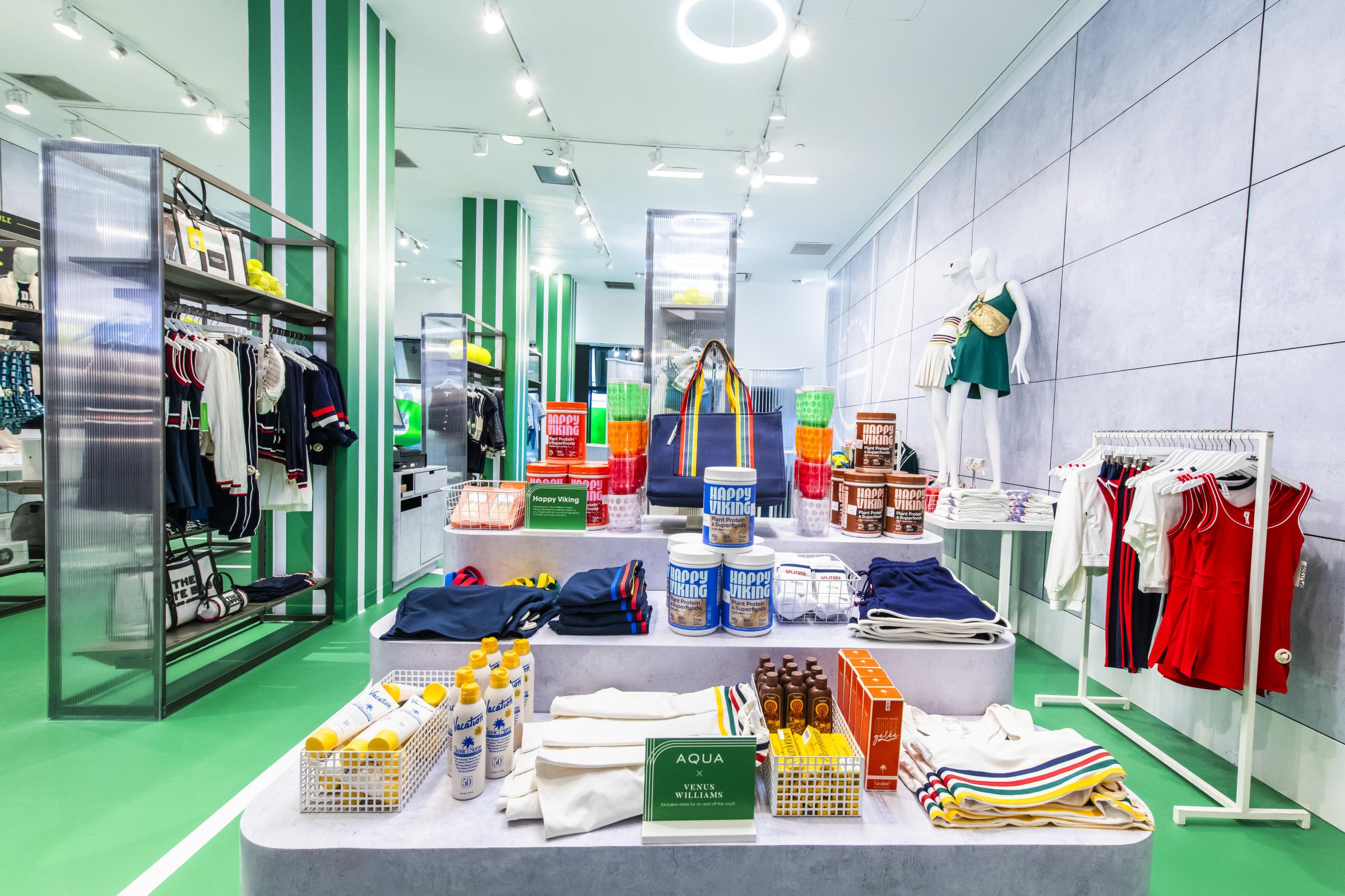 A retail store display with swimwear, accessories, and related products, including a mannequin dressed in a green outfit with a beige bag, colorful towels, and various bottles and cans of product packaging, on a modern, well-lit store interior with g