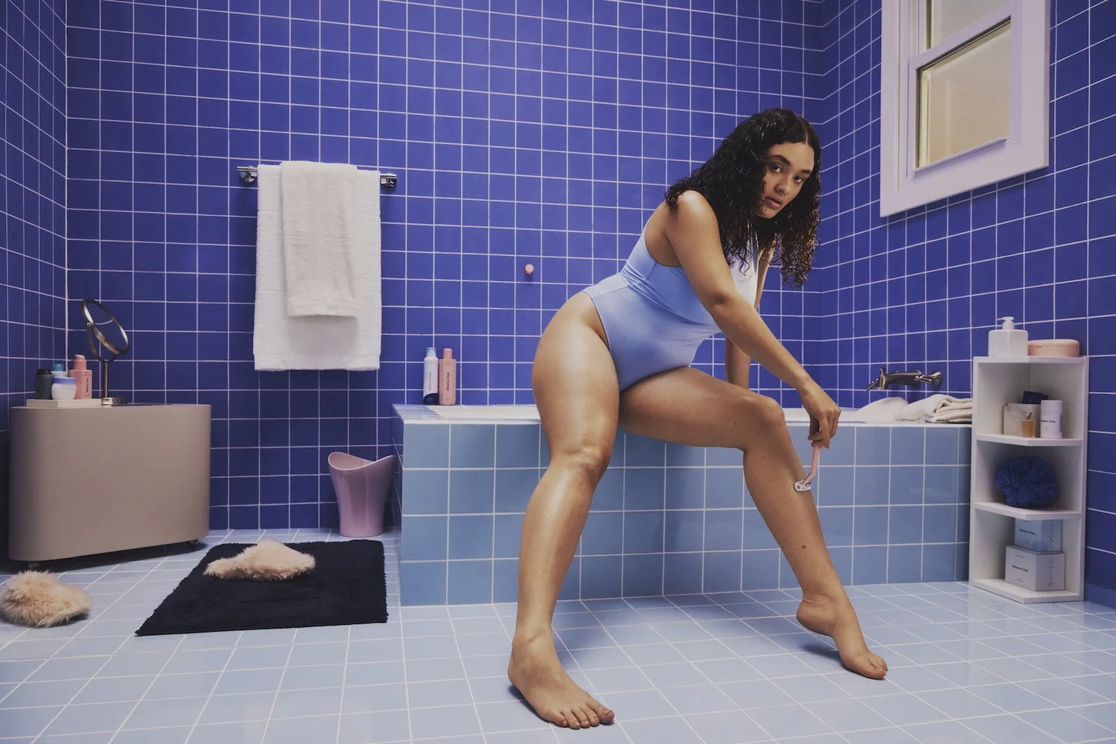 A woman in a light blue swimsuit is in a blue tiled bathroom, squatting and using a razor on her leg. The bathroom has a window, a white towel, and shelves with toiletries.