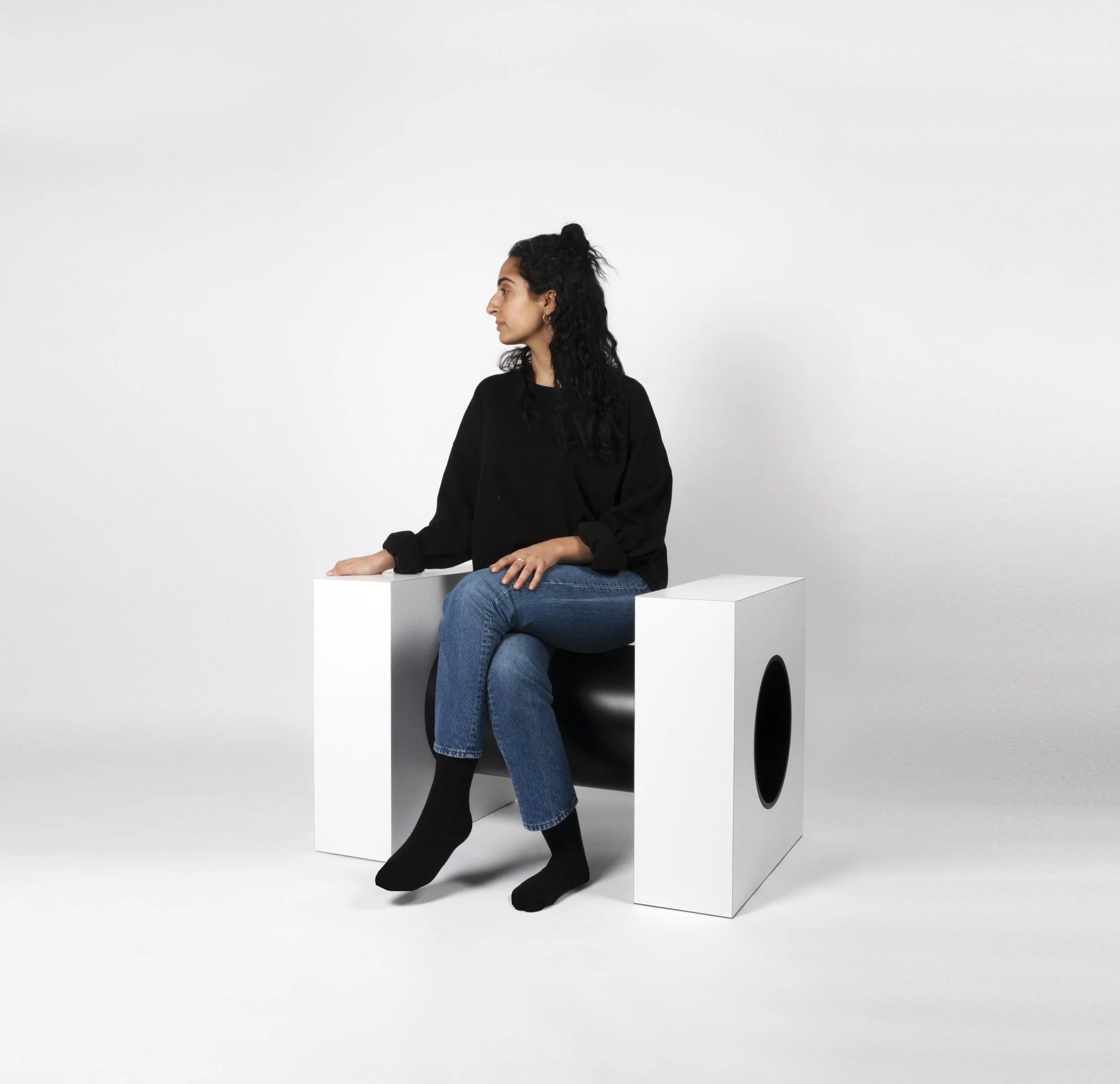 A woman with curly dark hair sitting on a black chair, wearing a black sweater, blue jeans, and black socks, next to two white rectangular blocks with black oval cutouts in a minimal studio setting.