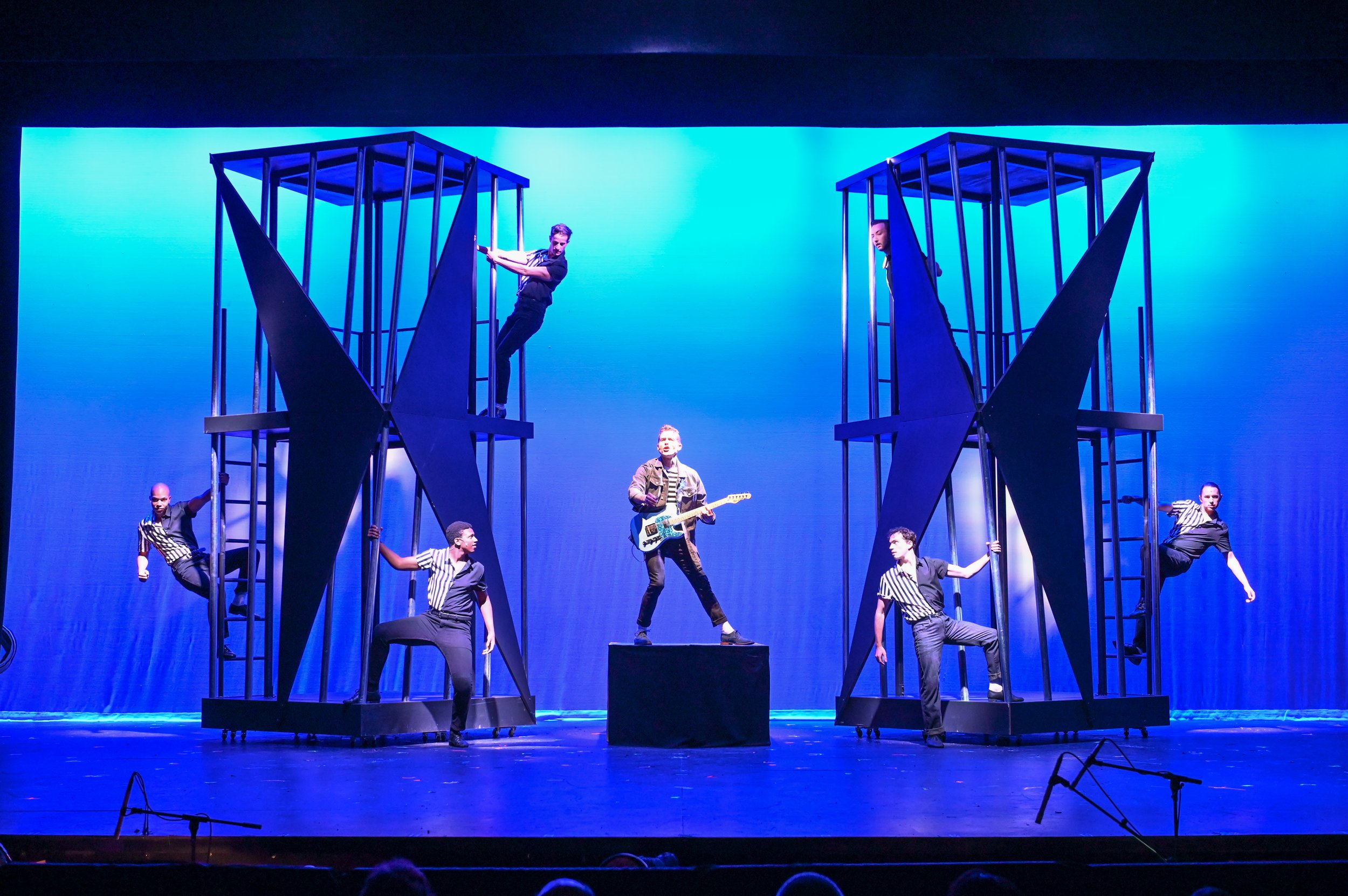 Stage performance with six performers, two on each of two tall black structures, one in the center holding an electric guitar, and two performers at the bottom of each structure dressed in black and white striped shirts, with a blue background.