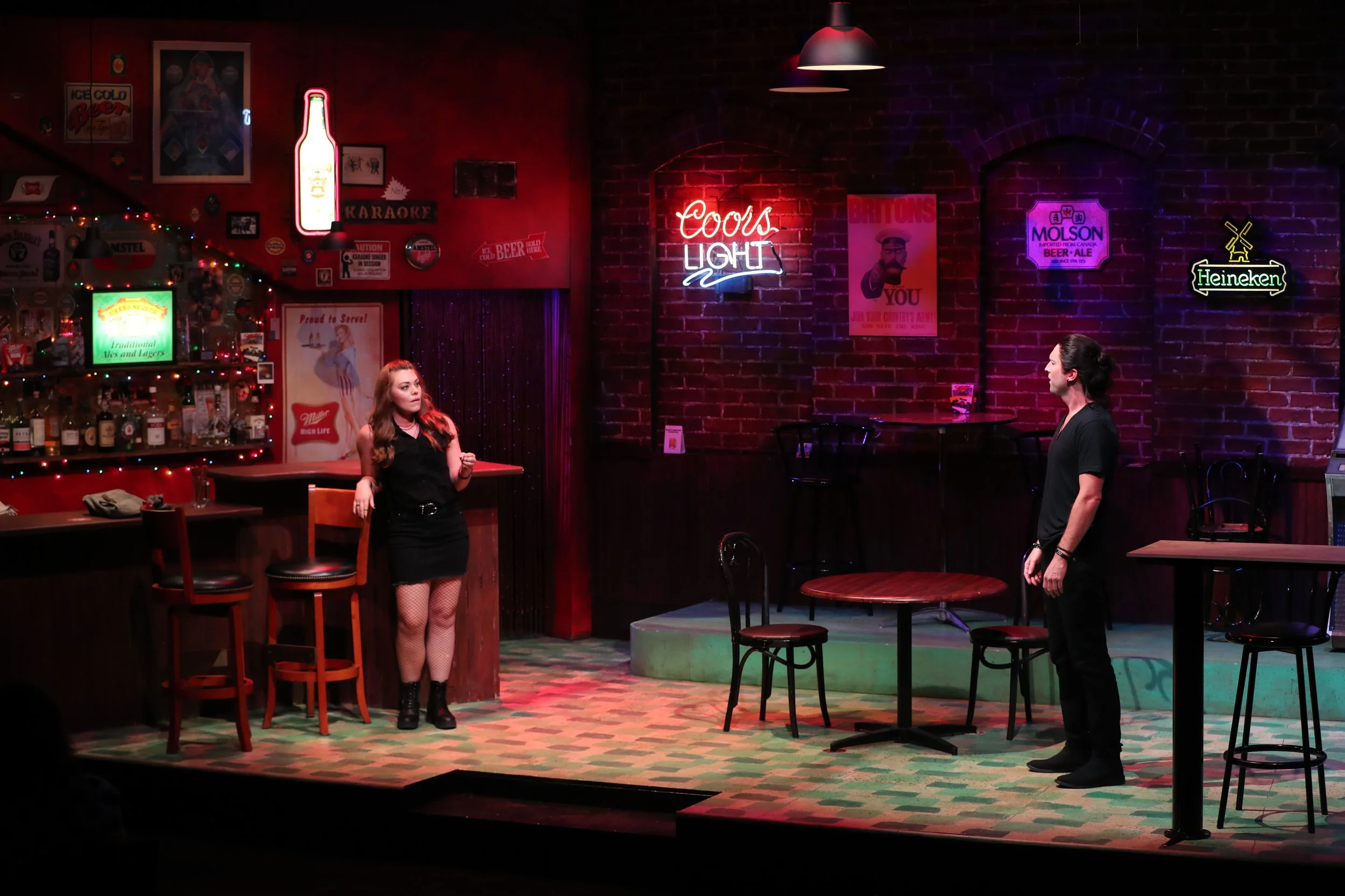 Scene from a comedic play or performance set in a bar with two actors, a woman on the left near the bar counter and a man on the right standing, surrounded by neon signs and bar memorabilia.