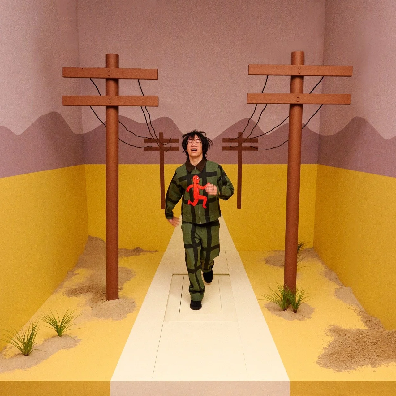 Person running on a white pathway, wearing a green and black striped outfit with a red figure design, in a room with yellow and pink walls, and painted mountains and power lines in the background.