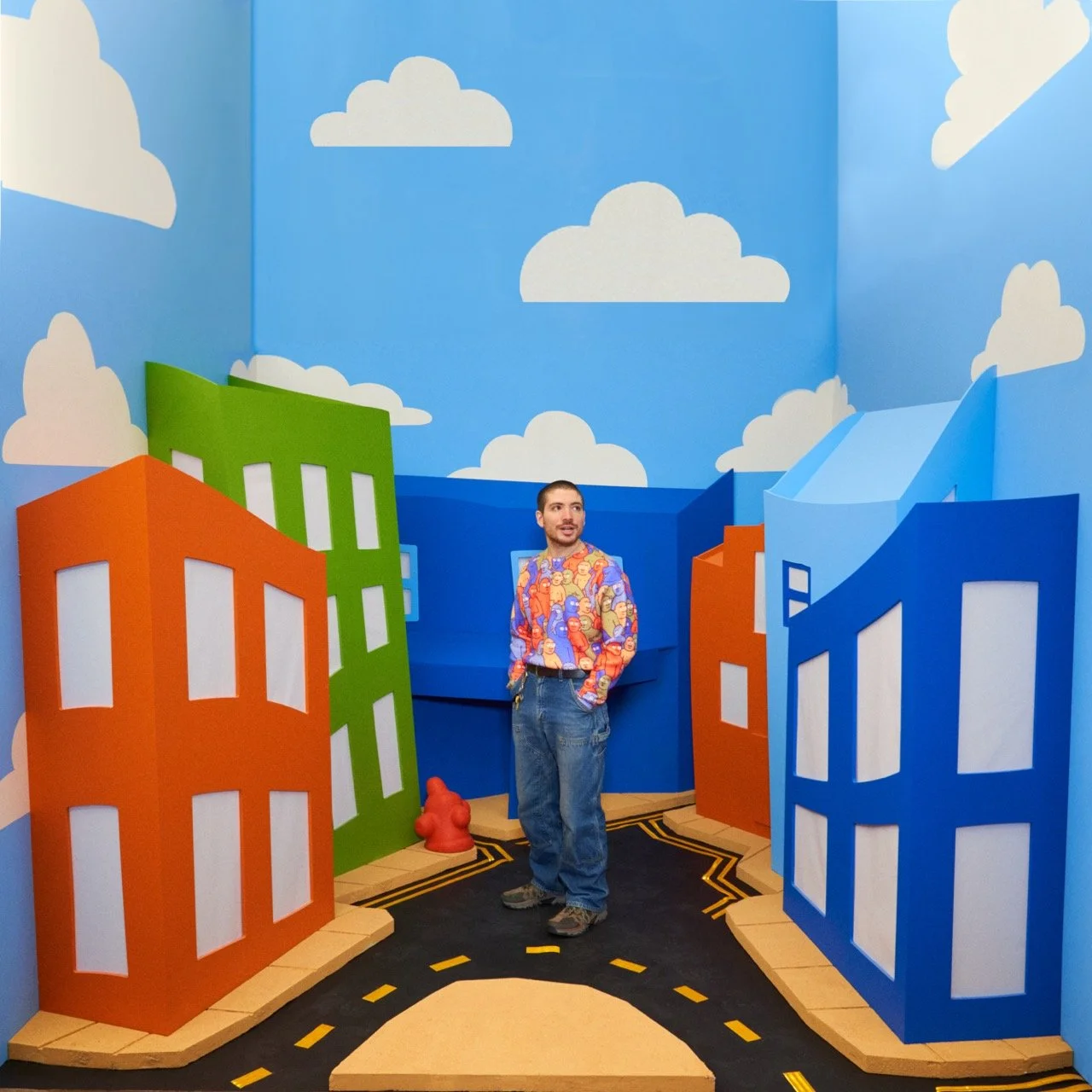 Person standing in a colorful, cartoon-style city scene with orange, green, and blue buildings, a road, and painted clouds on the wall.