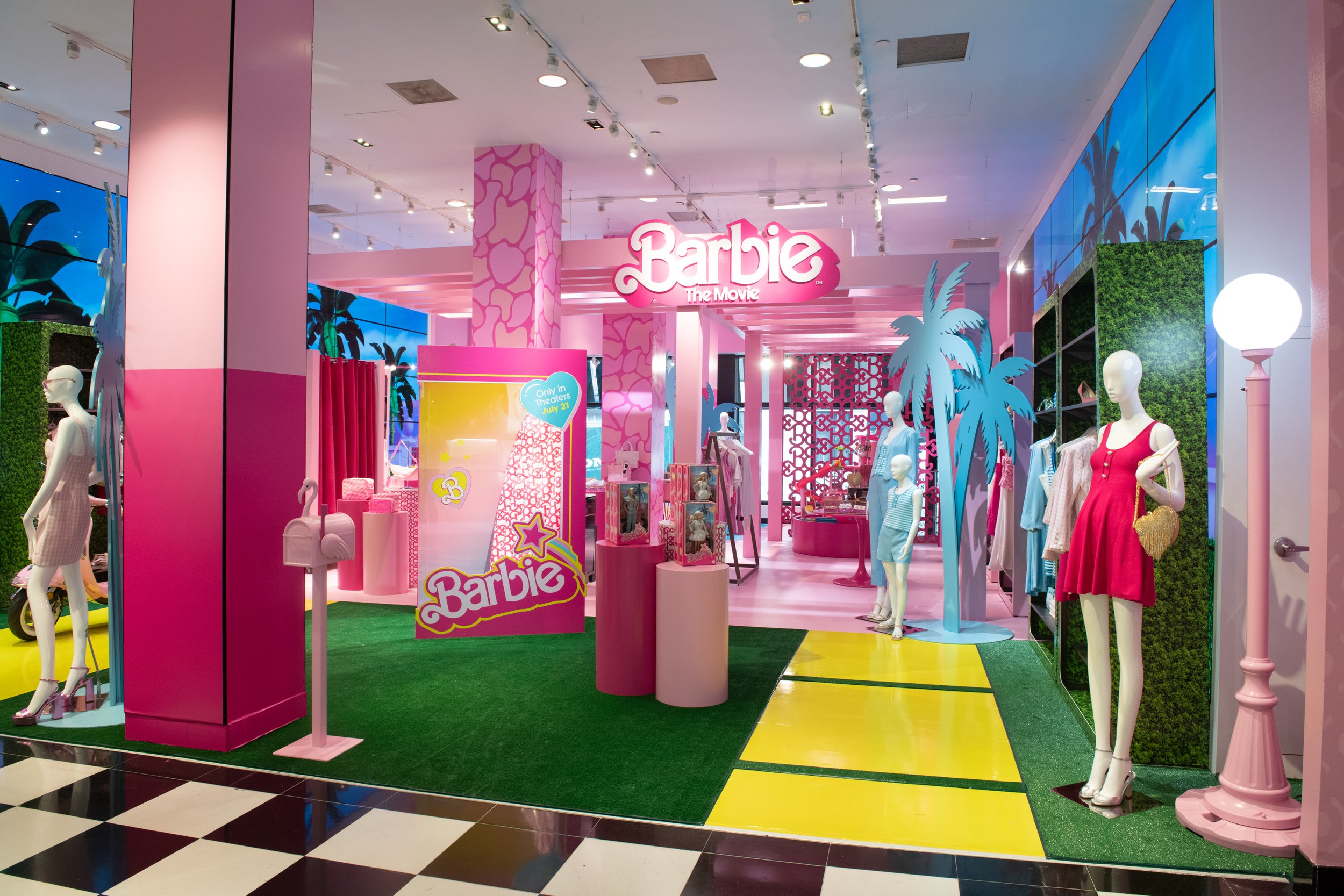 Interior of a Barbie-themed store with pink, blue, and yellow decor. Mannequins display various outfits, and there are palm tree decorations and a sign that reads 'Barbie The Movie.'