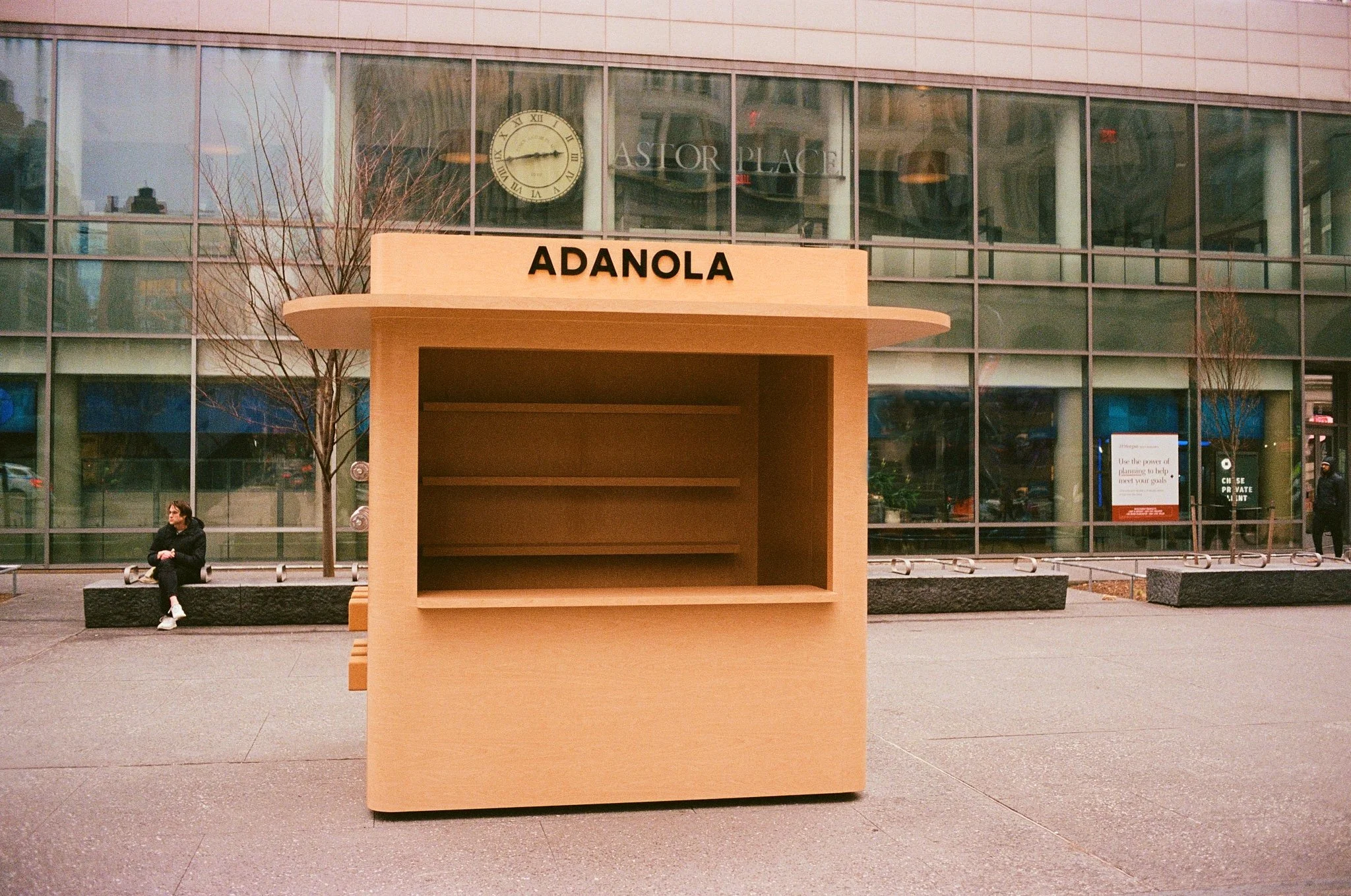 A customer engagement popup made out of light wood standalone kiosk with the word 'ADANOLA' on top, situated on a city sidewalk in front of a glass building.