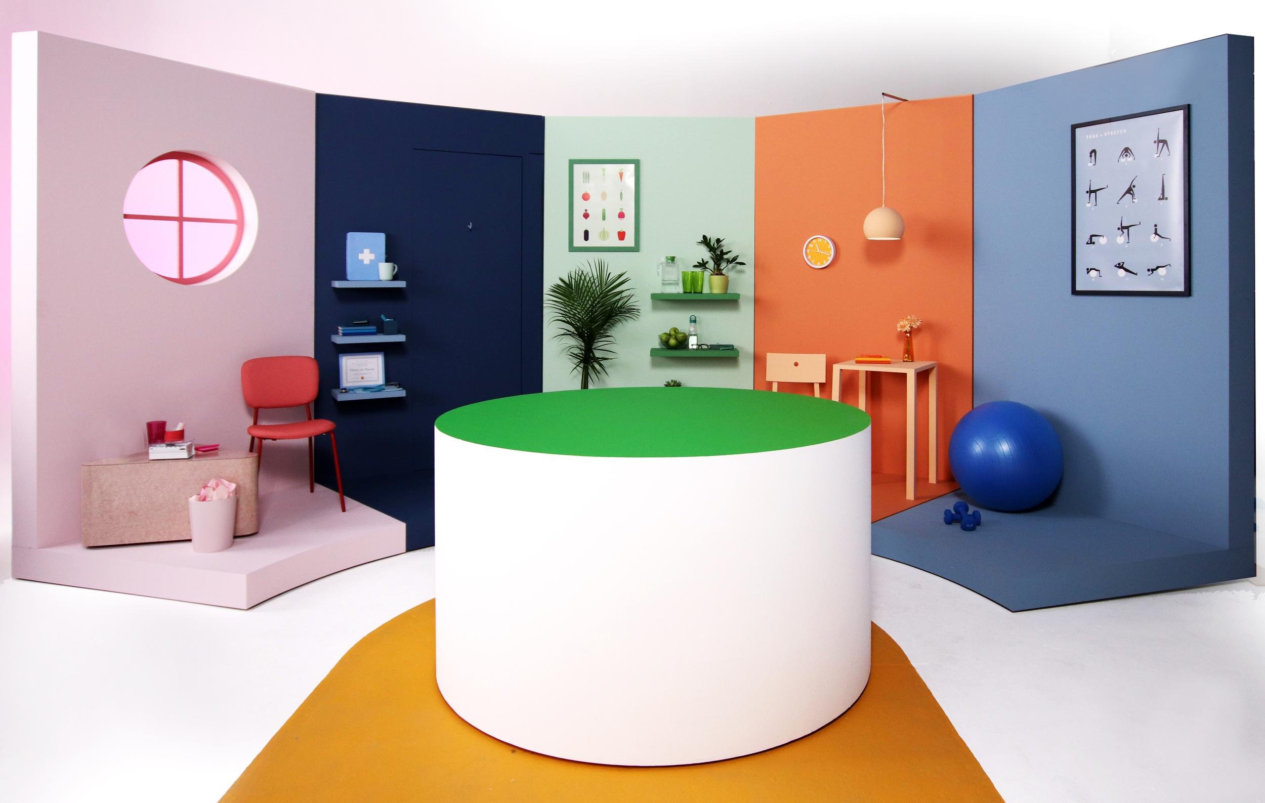 Colorful modern room with pastel walls, large green and white circular platform in the center, blue exercise ball, and framed artwork. Decor includes shelves with plants and books, a small table with a flower vase, and wall clocks.