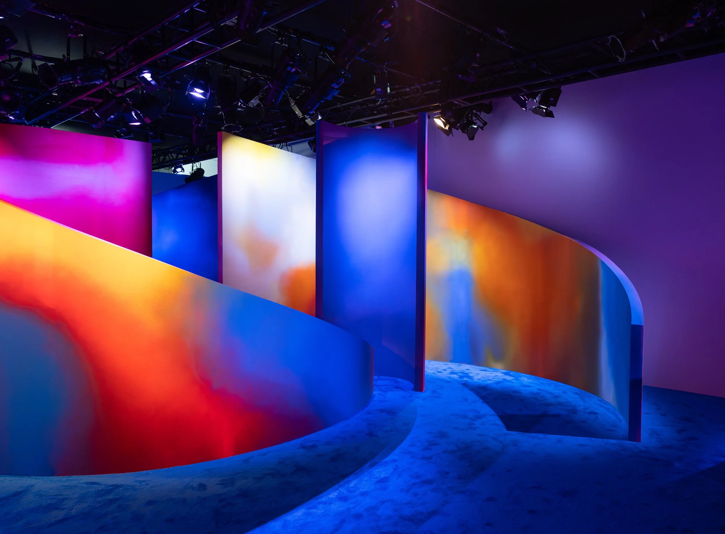 Colorful abstract art installation with flowing curved walls in vibrant pink, yellow, blue, and orange hues, set in a dark room with overhead lighting.