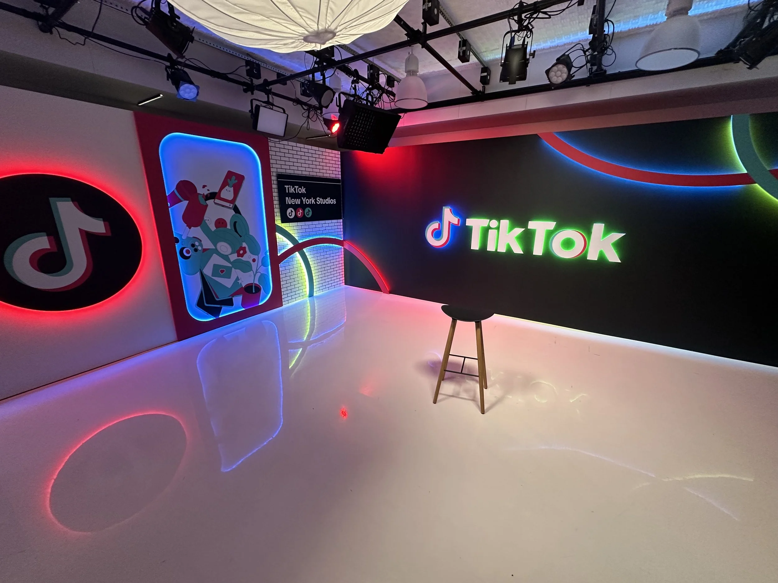 A brightly lit TikTok studio with colorful neon signs and wall art, including the TikTok logo and a graphic of a person with a phone, with a stool in the center.