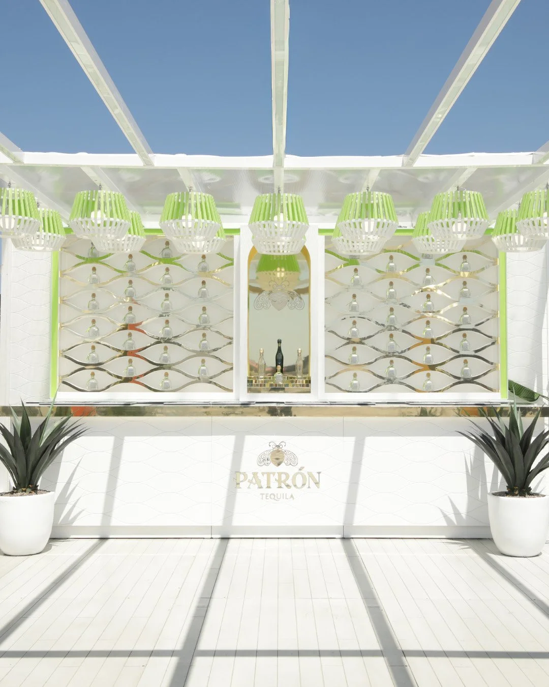 Modern outdoor bar with bottles of Patron tequila, decorative lighting, and potted plants, under a clear sky.