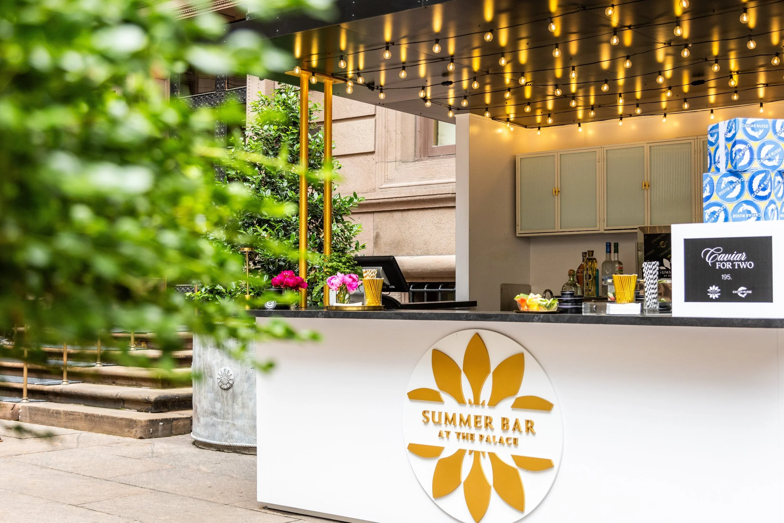 Outdoor bar station with string lights, flowers, and a sign that reads "Summer Bar at the Palace".