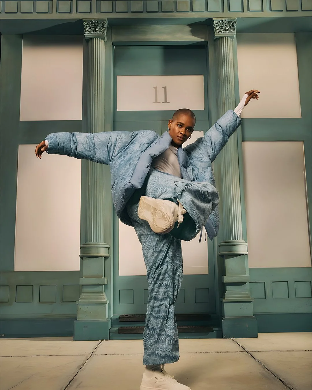 Person in blue patterned outfit striking a pose with one leg lifted, standing in front of a light teal building with columns and a sign numbered '11'.