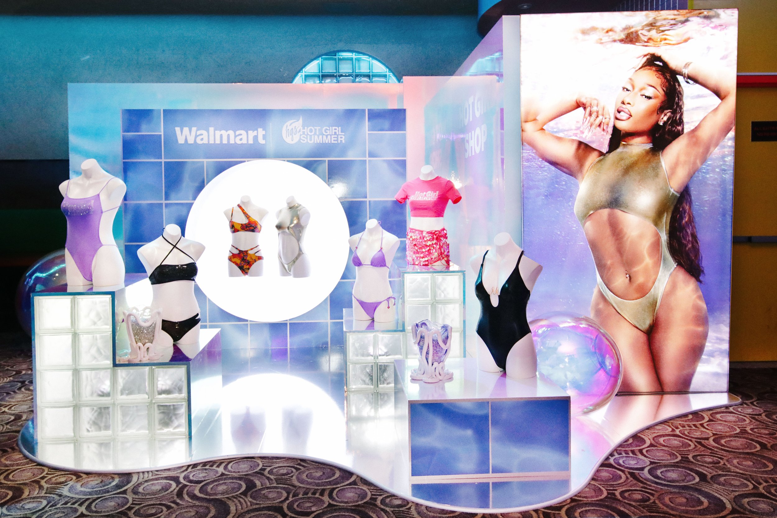 Display of mannequins showcasing swimwear and loungewear with a large poster of a woman in a metallic swimsuit in the background. The display features various bikini and one-piece swimsuits in different colors and patterns, set on illuminated platfor