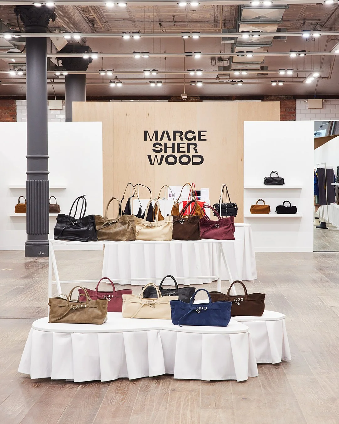 Display of handbags arranged on white tiered stands in a store with a sign reading 'MARG SHE R WOOD' in the background.