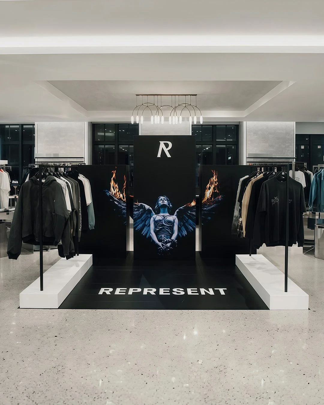 Display of clothing in a retail store with a large central poster featuring a woman with dark wings and flames, black and white racks of clothing on sides, modern interior design.