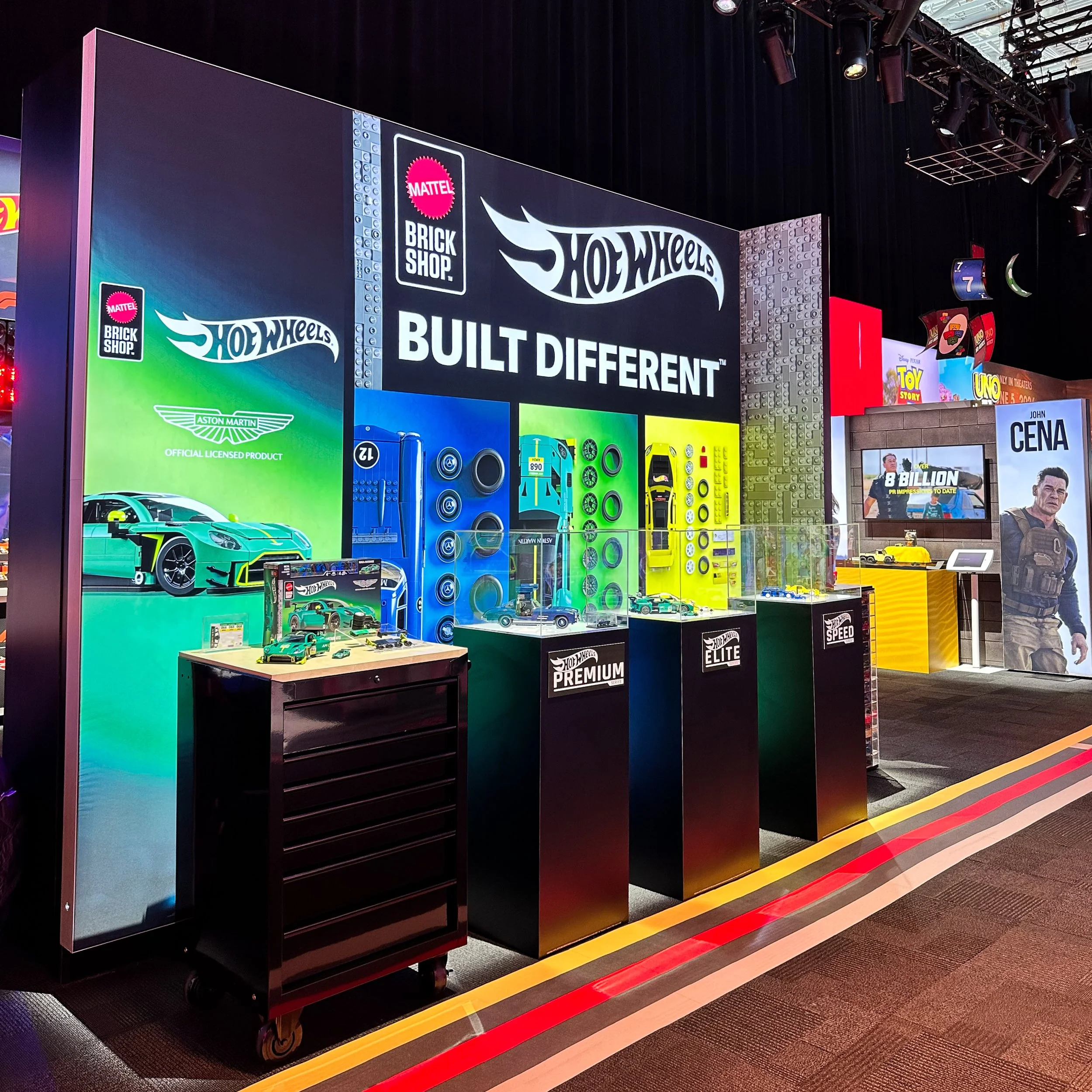 Hot Wheels display at an event featuring various toy cars, branded products, and promotional banners with the Hot Wheels logo and slogans such as 'Built Different' and 'Brick Shop', and several large posters including Aston Martin and John Cena image