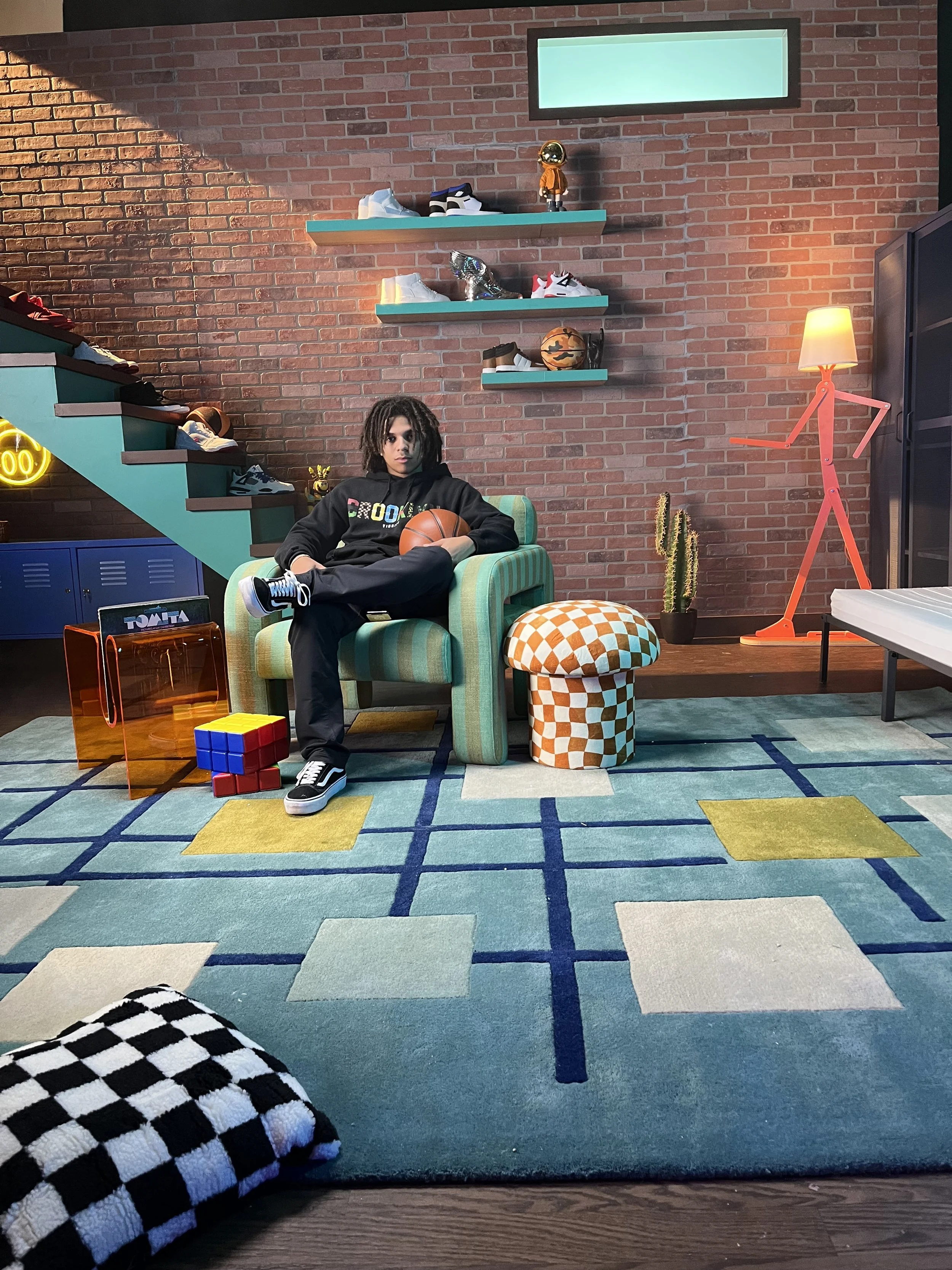 Teen boy with dreadlocks sitting on a green striped armchair, holding a basketball, with shoes, a Rubik's cube, and a translucent orange folder on the floor nearby, in a modern room with a brick wall, floating shelves with sneakers, a cactus, a hat, 
