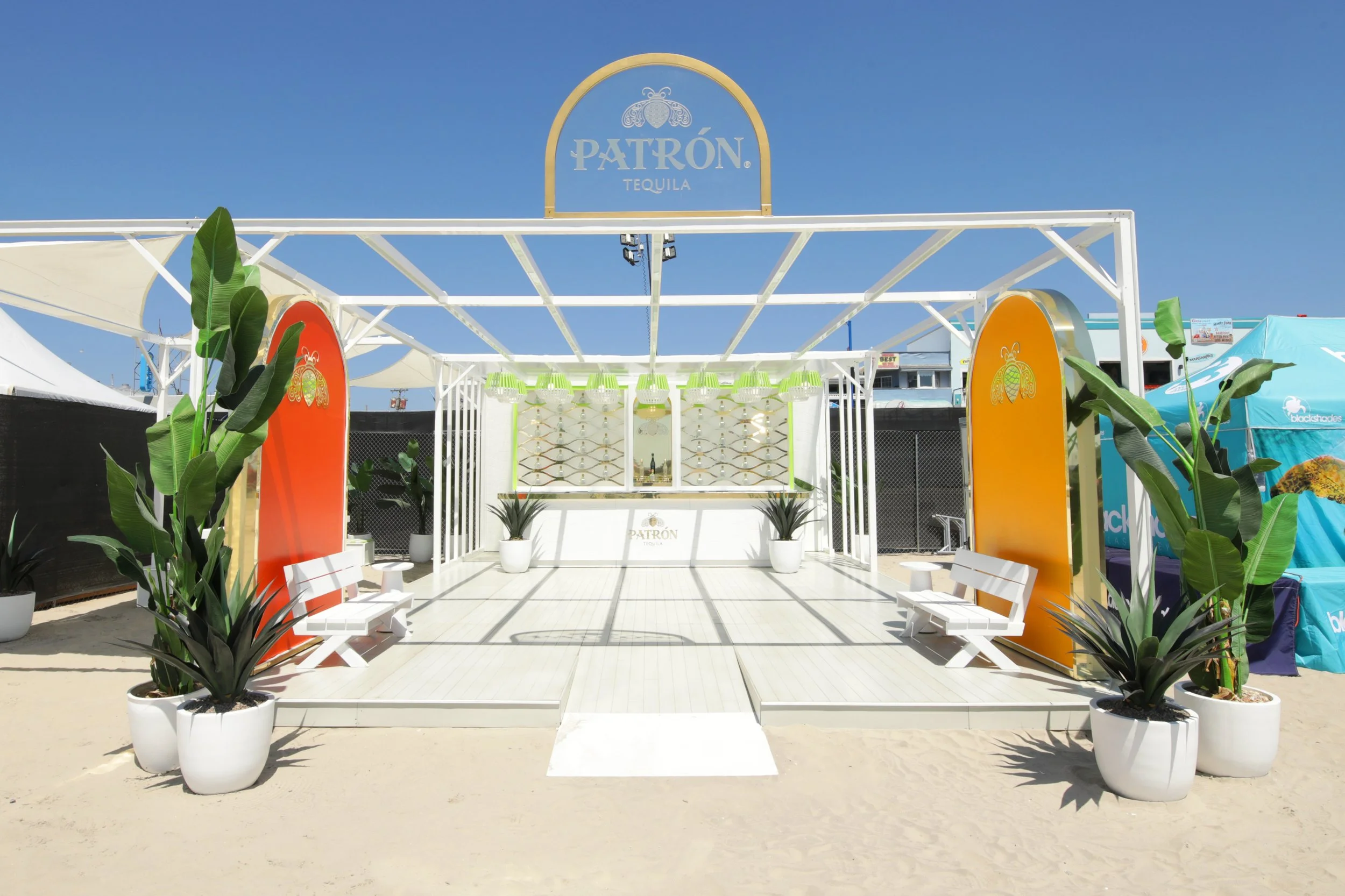 Beach bar setup with white patio furniture, potted plants, and a sign that reads 'Patrón Tequila' under a clear blue sky.