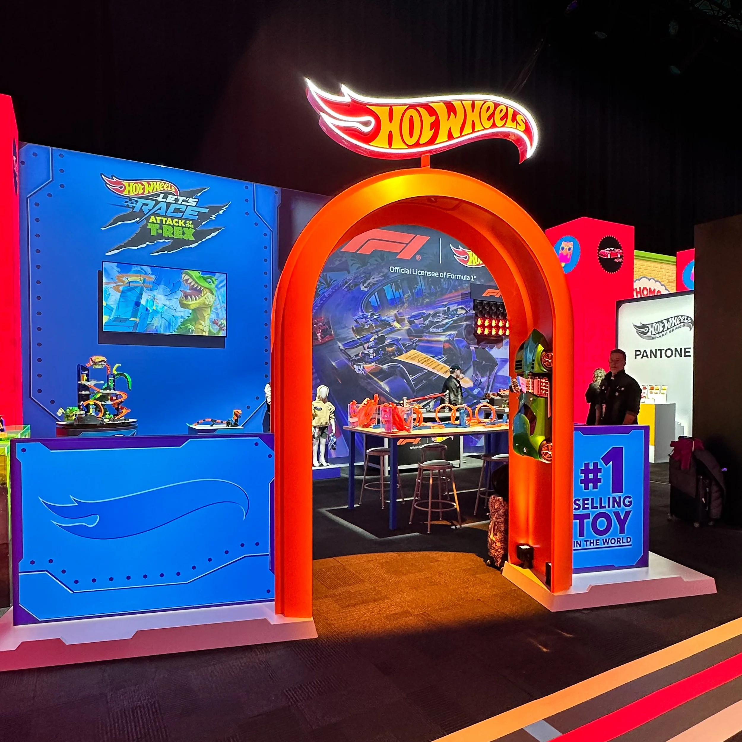 Colorful Hot Wheels toy display at an event or showroom, featuring a large illuminated Hot Wheels sign, a blue display booth with toys and a screen, and a sign stating '#1 Selling Toy in the World'.