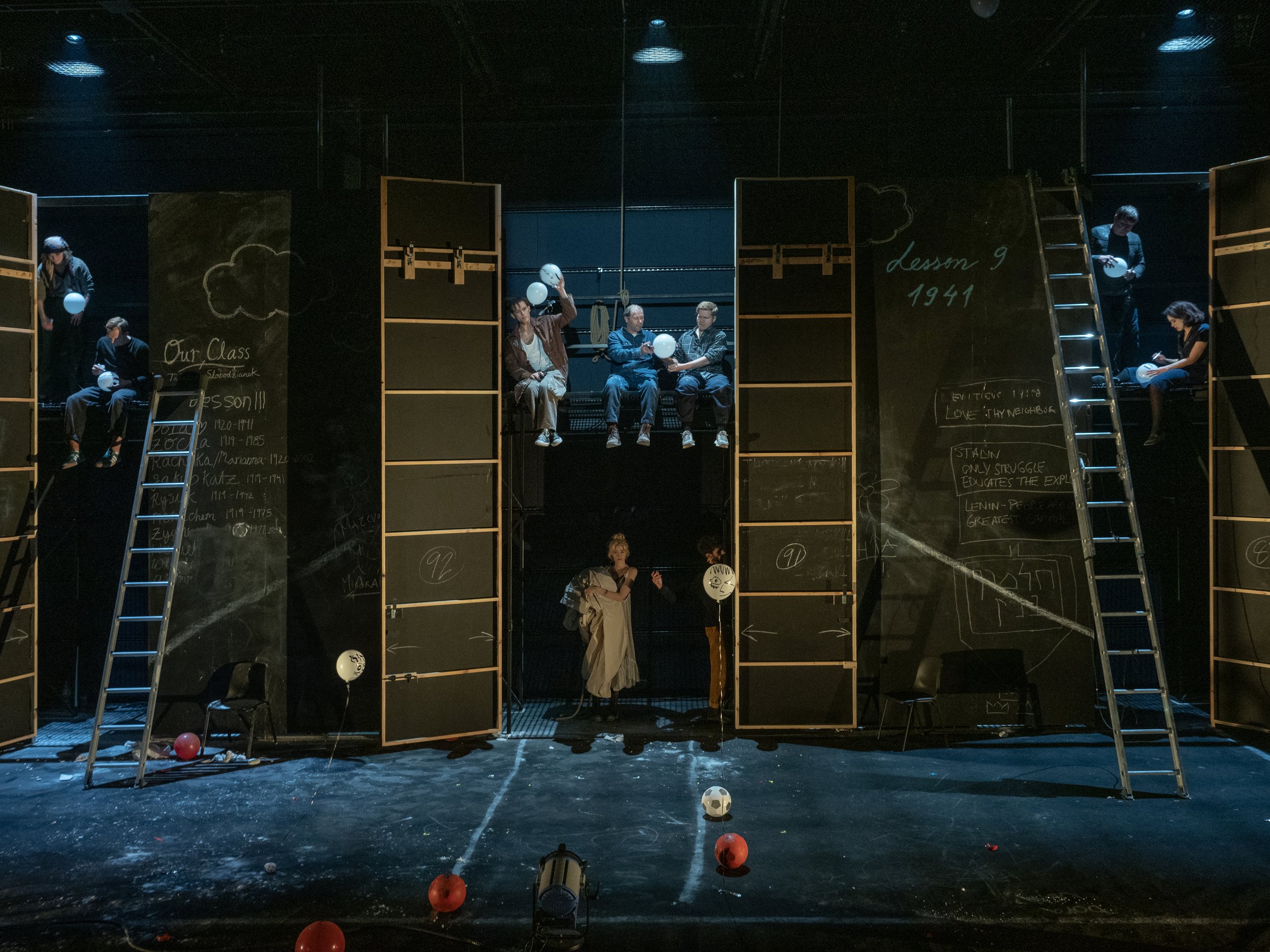Stage scene depicting students sitting on elevated platforms, some holding balloons, with chalkboards and ladders, and stage lights overhead.