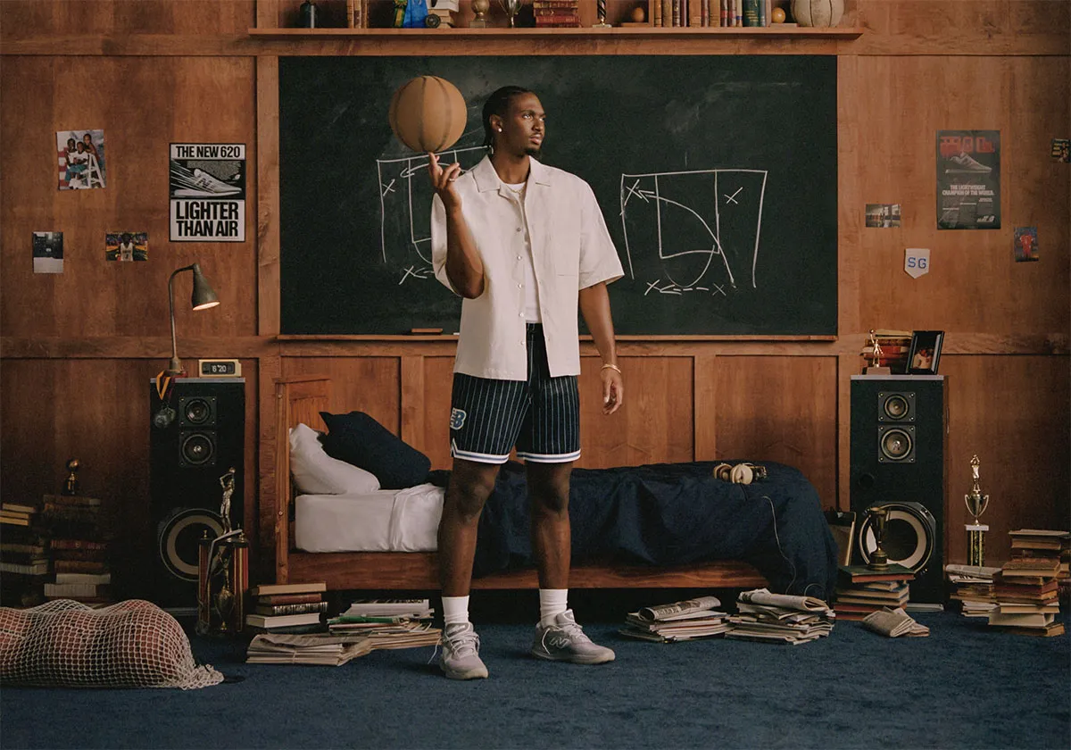 A man in a basketball jersey and shorts standing in a bedroom, spinning a basketball on his finger, with chalk-drawn basketball plays on a blackboard behind him.