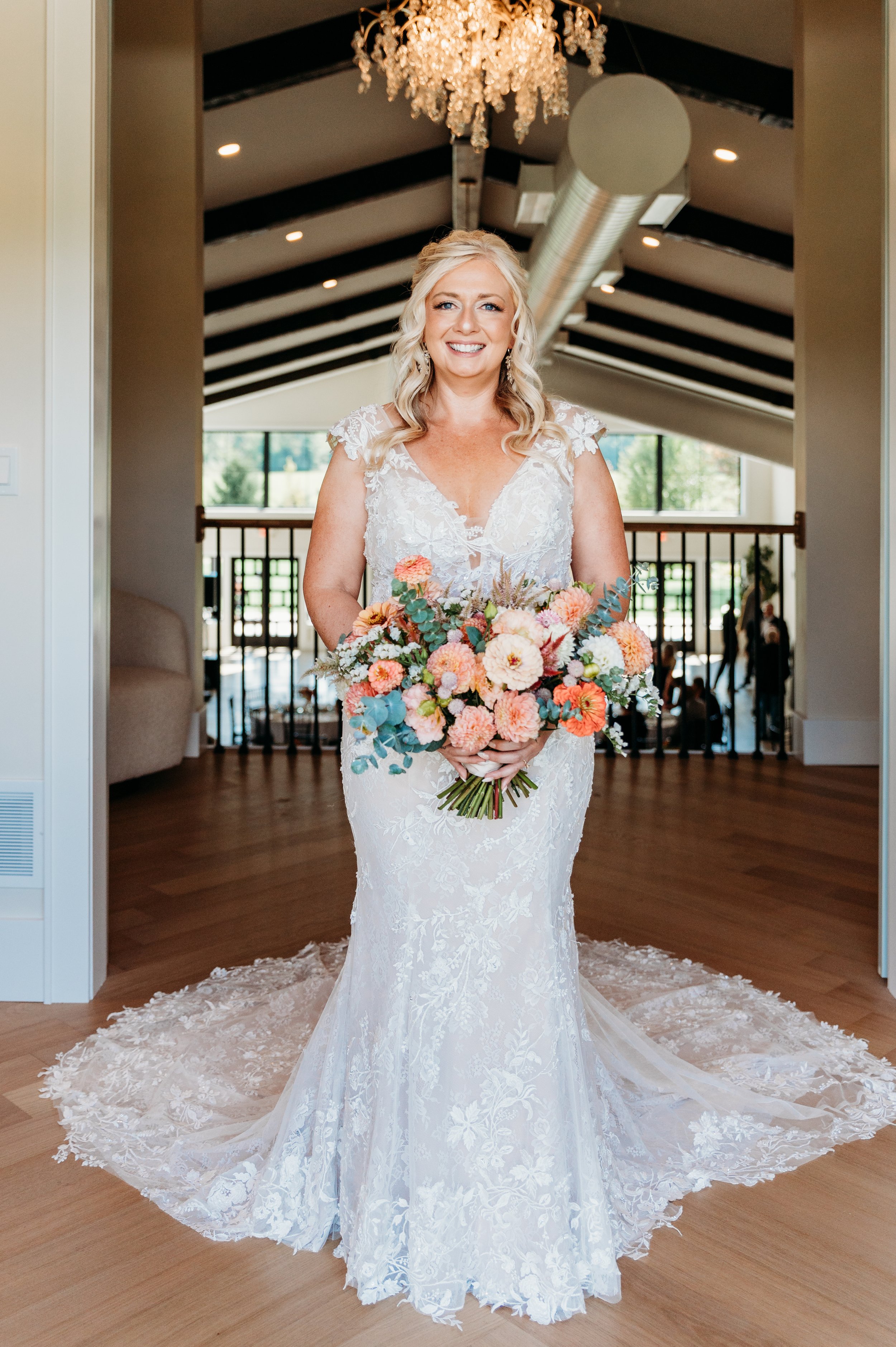 A bride in a white lace wedding dress holding a large bouquet of pink, peach, and white flowers on her wedding day, standing indoors with a chandelier and large windows in the background.