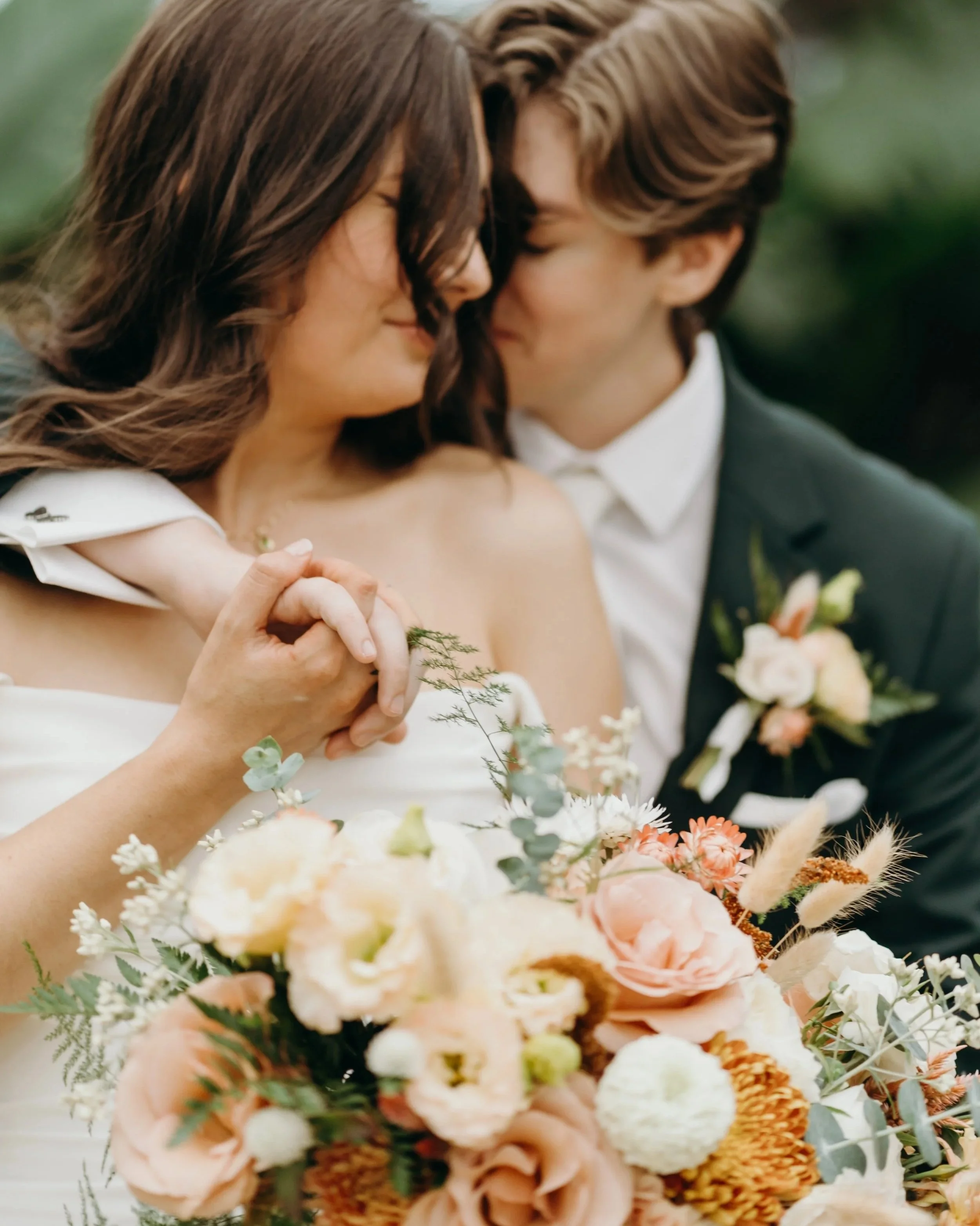 A bride and groom close together on their wedding day, the bride holding a bouquet of flowers, and they are gently touching foreheads with eyes closed.