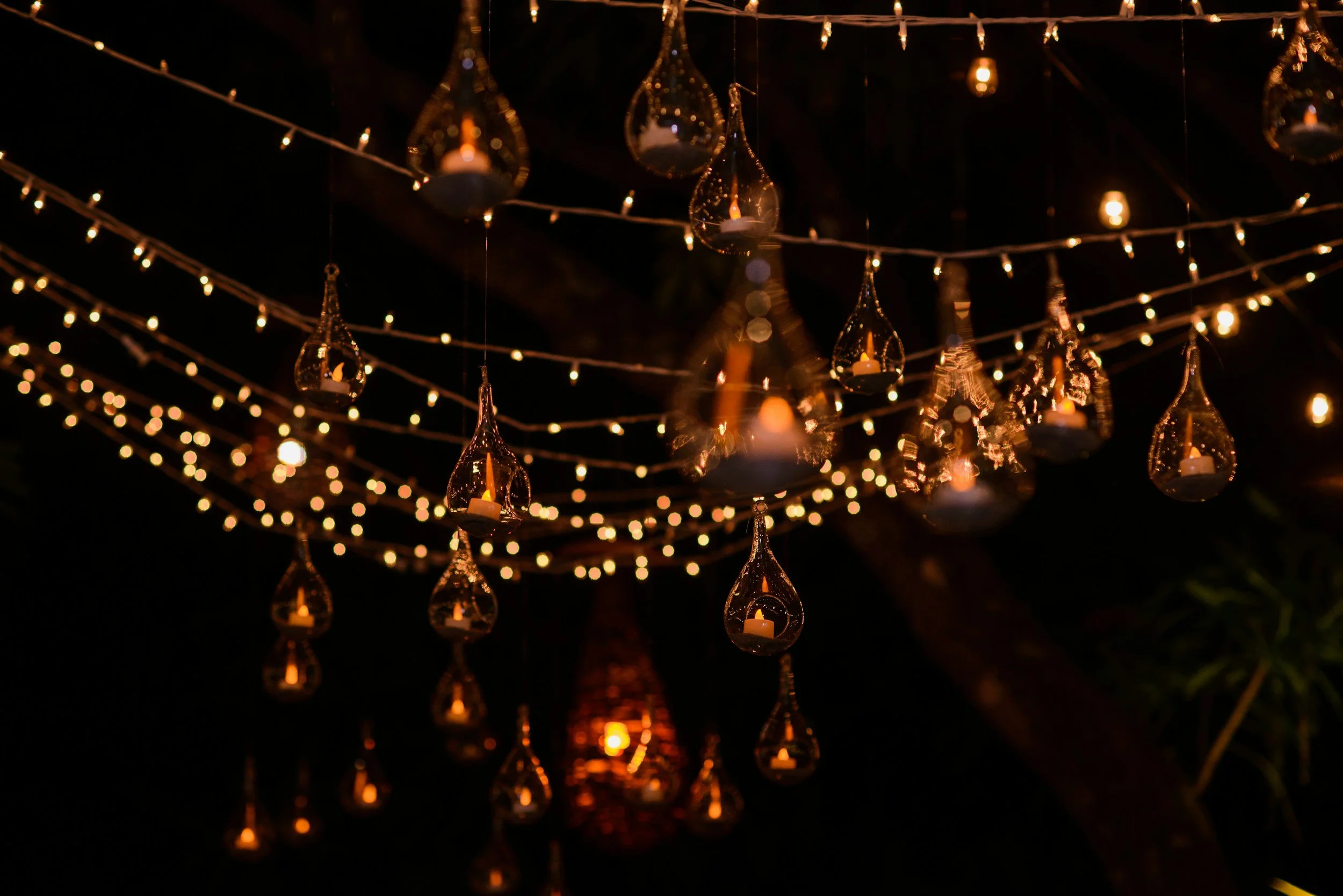 String of hanging glass lanterns with candles inside, illuminated and glowing against a dark night sky, decorated with string lights for a festive or romantic outdoor setting.