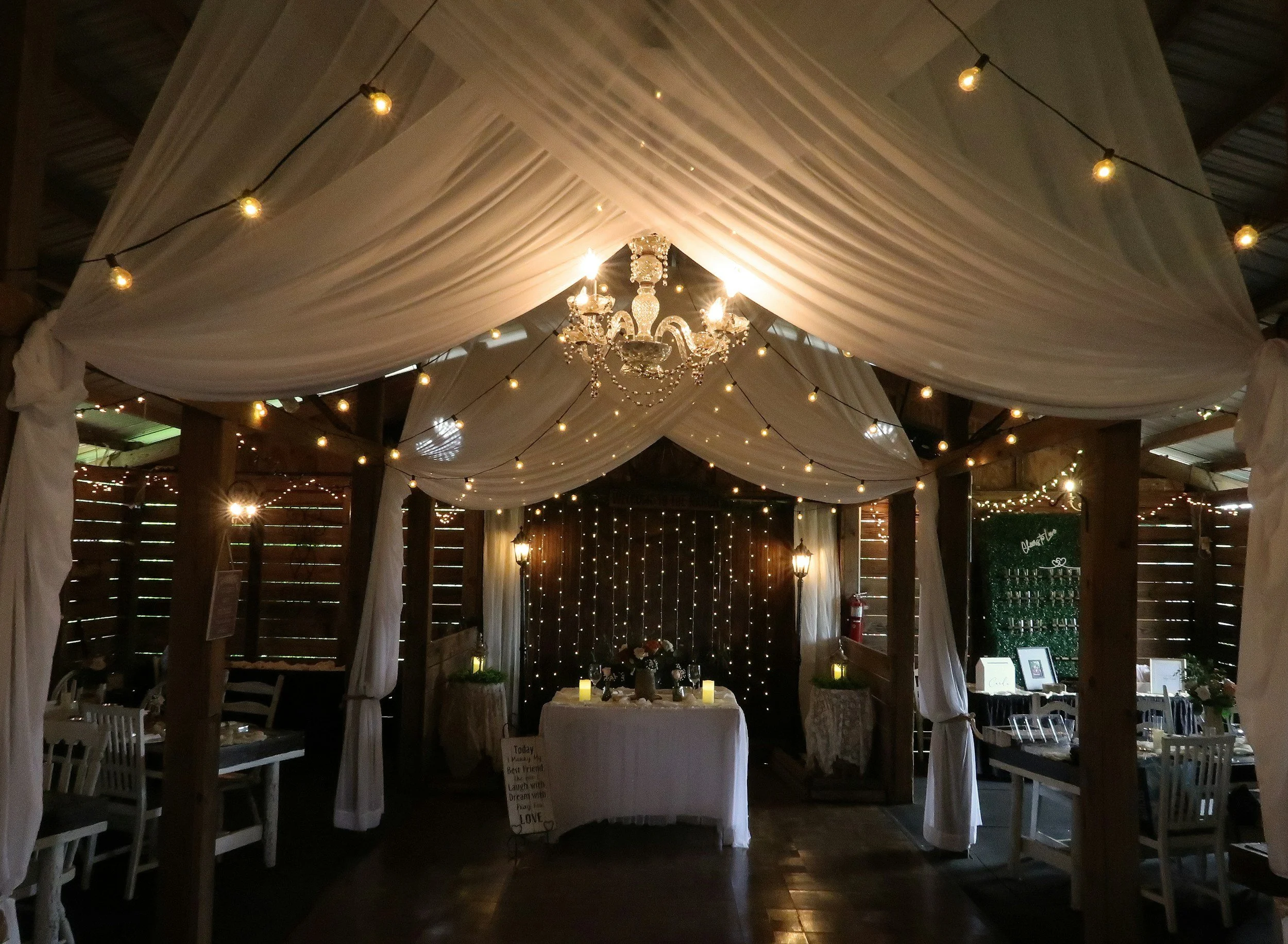 A decorated event space with draped white fabric ceiling, ambient string lights, and a chandelier, featuring tables and chairs arranged for a celebration.